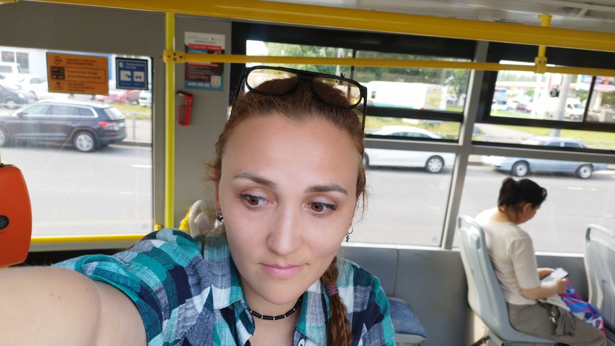 A woman with auburn hair, wearing a blue and green plaid shirt and a black necklace, sits on a bus. Her sunglasses are perched on her head, and she looks down and to the side with a pensive expression. Another woman with dark hair is seated behind her, looking at her phone. Outside the bus window, cars are visible on a street, suggesting it is daytime. The scene is set within a bus in Saint Petersburg, Russia. There is some visible text on signage inside the bus, including a yellow sign with Cyrillic text and a blue sign with Cyrillic text.