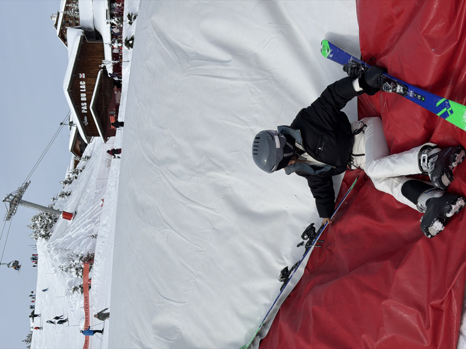 The image captures a snowy mountain ski resort scene, specifically identified by the "PAS DU LAC" sign, indicating the Pas du Lac area within Méribel, part of Les Allues, France. In the foreground, an individual is seated on a red inflatable safety barrier or cushion. This person is wearing a grey ski helmet with goggles, a black insulated jacket, white ski pants, and black ski boots. They are holding a pair of blue and green skis, one on each side, with their gloved hands resting on the ski bindings. A large white inflatable structure forms a backdrop directly behind the seated individual.
The midground features a wide, snow-covered ski slope where multiple skiers are visible, actively skiing or standing. A large metal ski lift pylon with a red base is positioned on the slope to the left. A red safety fence or netting runs across the lower part of the slope. To the right, a prominent multi-story wooden chalet-style building, marked with the sign "PAS DU LAC," stands with snow-covered roofs, suggesting a restaurant or lodge.
In the background, more snow-covered slopes rise, dotted with evergreen trees. A chairlift system with moving chairs and additional skiers is visible further up the mountain. The sky is uniformly overcast. The scene depicts a busy winter sports environment.