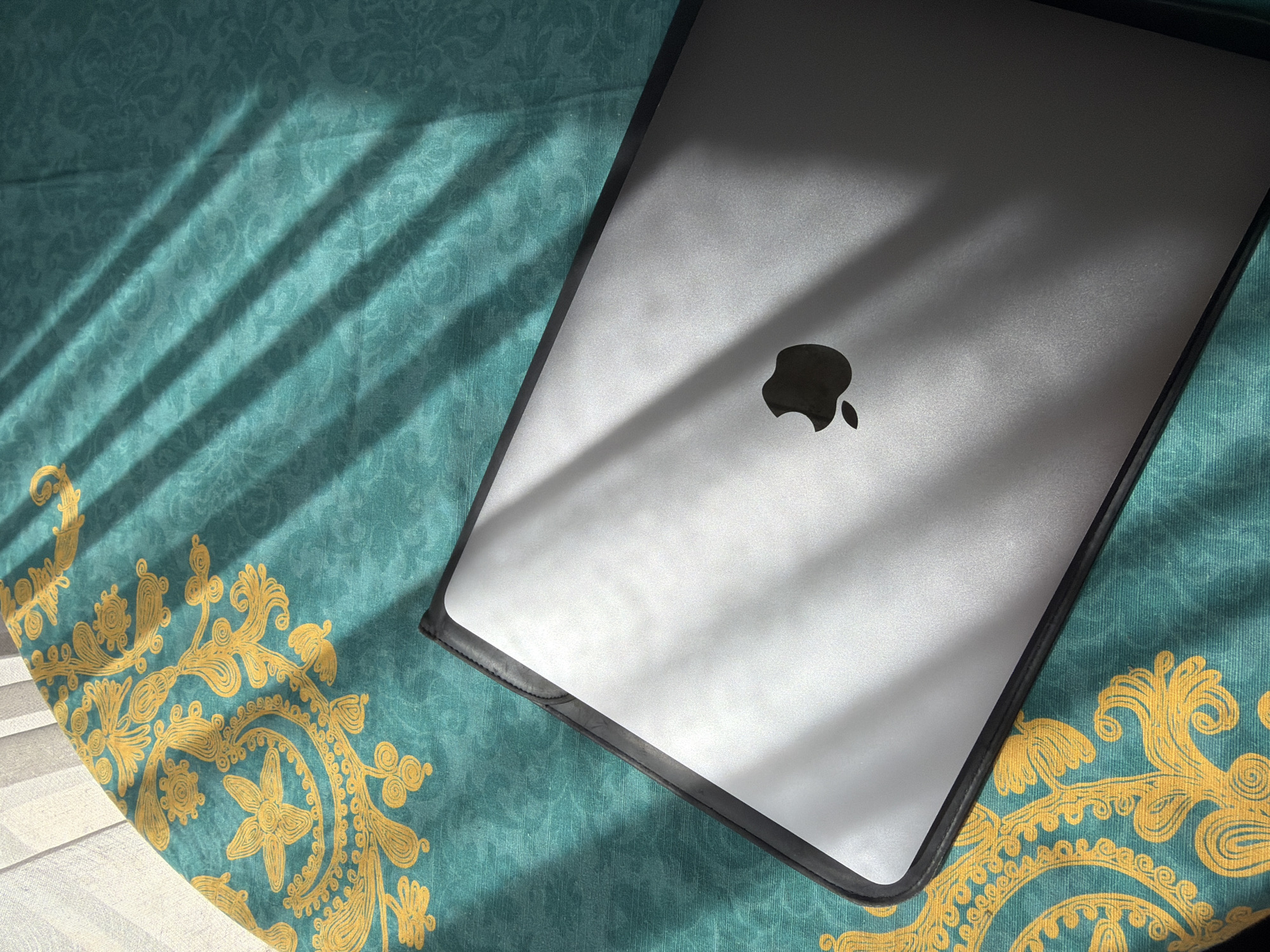 A silver Apple laptop sits on a teal patterned fabric with gold designs, bathed in the dappled light from a window. The setting is Tbilisi, Georgia.