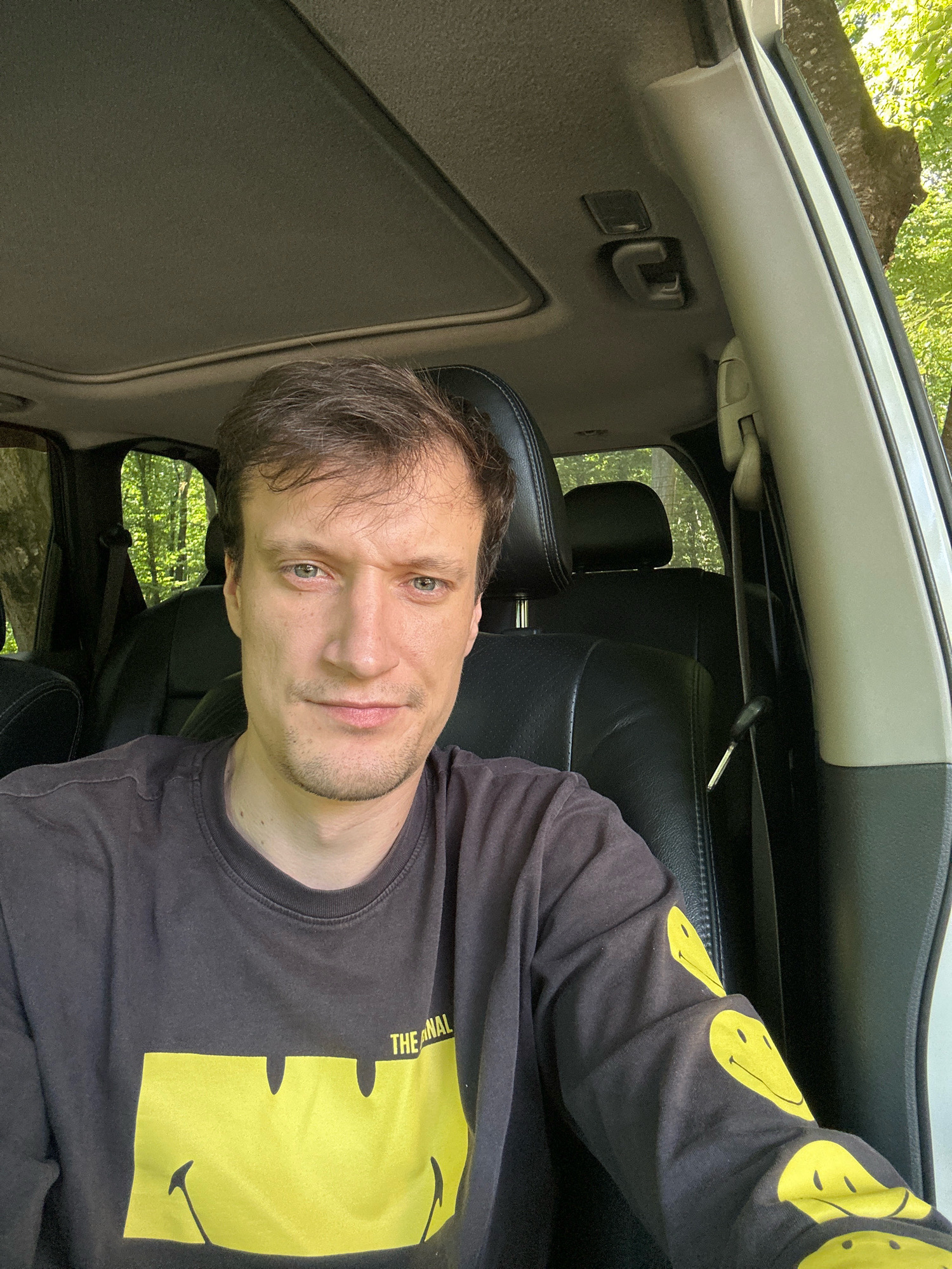 A man is sitting in the driver's seat of a vehicle. The vehicle appears to be parked in a wooded area, as evidenced by the trees visible through the windows. The man is wearing a dark long-sleeved shirt with a large yellow smiley face graphic on the front and smaller smiley face graphics on the sleeves. He is looking at the camera. The interior of the car is visible, including the dashboard, steering wheel, and seats. The image is a selfie, taken from a slightly high angle. The man's facial expression is neutral. The lighting suggests it is daytime. There is no information about the specific location within the city of Shatava, Ukraine.