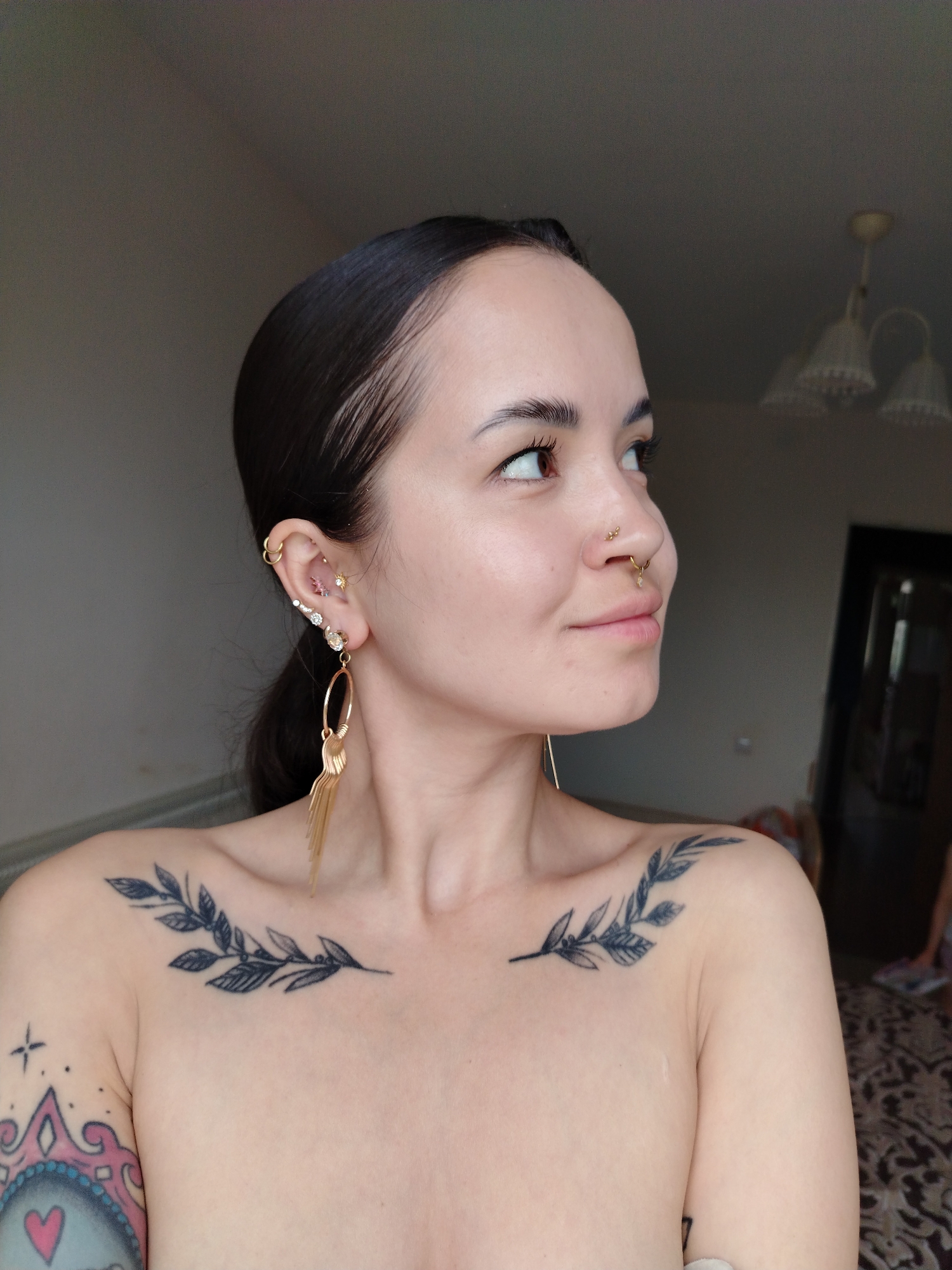 A young woman poses for a close-up indoor photograph. She has dark hair pulled back, fair skin, and numerous piercings in her ear and nose. She wears gold earrings. Black leafy branch tattoos decorate her upper chest, and another tattoo is visible on her left upper arm. The background includes a room with a chandelier and a doorway. The photo location is specified as Noviy, Russia.
