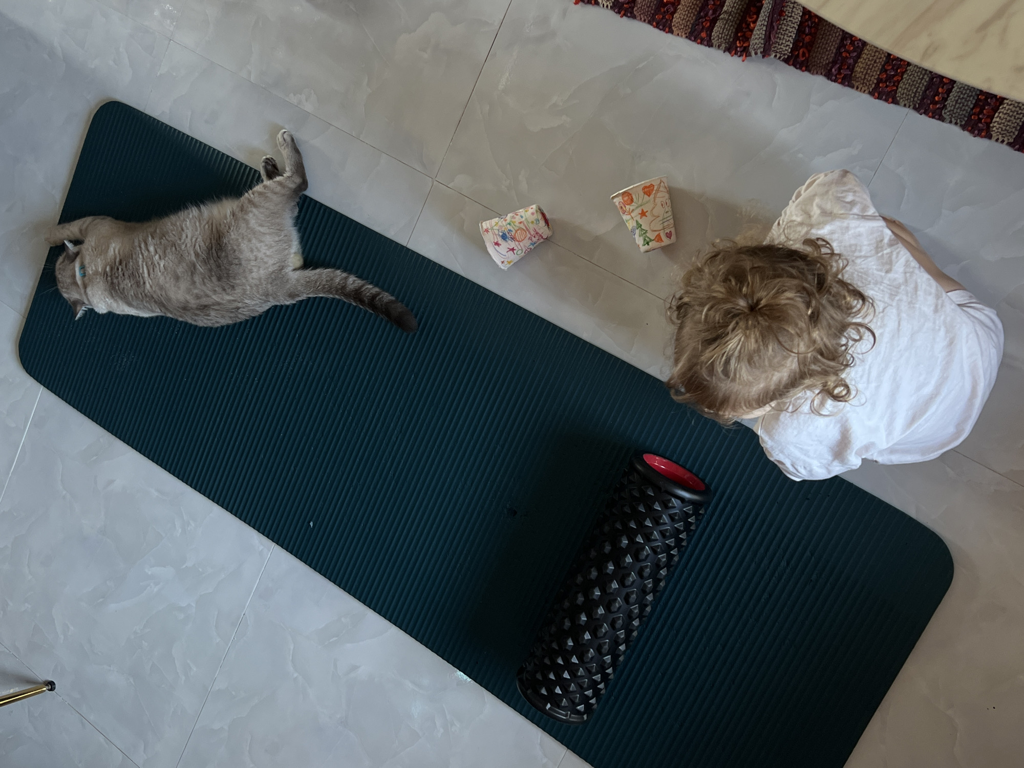 An overhead, eye-level shot captures a gray cat sprawled on a dark green yoga mat, its body extending horizontally. To the left of the mat, a black foam roller with red end caps rests on the mat. A young child, wearing a white t-shirt and with curly blond hair, is seated on the floor to the right of the mat, facing away from the camera. Scattered on the floor between the child and the yoga mat are two small, colorful paper cups. The floor is tiled with a light gray, marble-like pattern. A woven rug with red, gray, and purple stripes is visible in the lower right corner, and a portion of a light-colored table is also present. The scene is indoors, with no discernible background or city details.