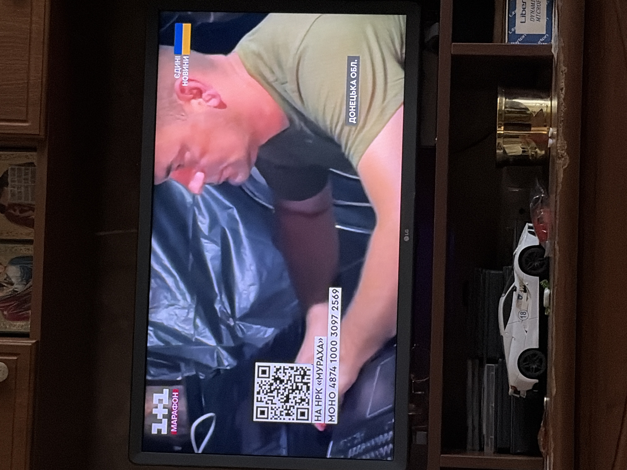 A television screen displays a news broadcast. The screen shows a man in a green t-shirt looking down and to his right. To the left of the screen, a QR code is visible, followed by text that reads "НА НРК «МУРАХА»" and a phone number. In the upper left corner of the screen, a logo for "1+1 МАРАФОН" is displayed. In the upper right corner, text reads "ЄДИНИ НОВИНИ" with the Ukrainian flag. Below the man's arm, text reads "ДОНЕЦЬКА ОБЛ.". The television is placed on a wooden shelf below. On the shelf, there is a white toy sports car with the number "18" on its side. To the right of the car, a gold cylindrical object and a box labeled "Liber" are visible. The background appears to be an indoor setting with wooden furniture.