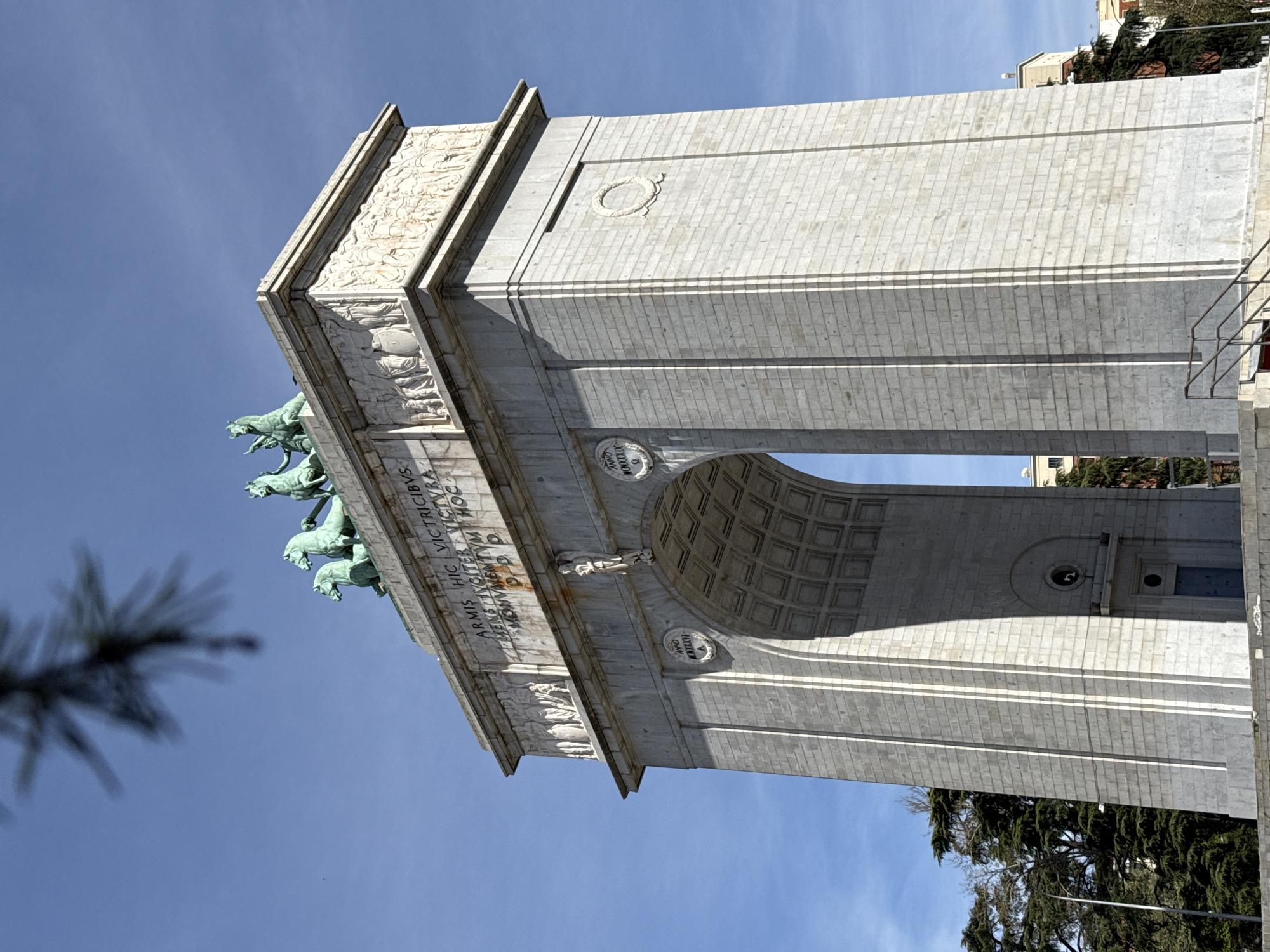 The image shows the Arco de la Victoria in Madrid, Spain. The monument is constructed from white stone and features a central archway. On top of the arch is a bronze quadriga with horses and a driver. The monument is decorated with carvings and inscriptions. The surrounding environment includes a blue sky and some trees.