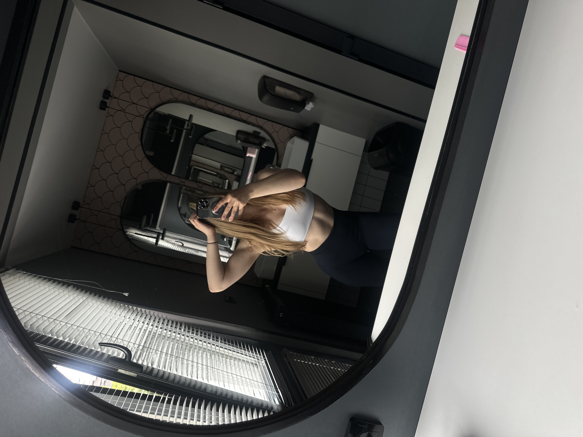 A young woman captures a mirror selfie in a modern restroom located in Ternopil, Ukraine. The woman, reflected from her chest to upper thighs, has long, light brown hair partially obscuring her face as she holds a smartphone (identified by its triple-lens camera setup) with her right hand. Her left hand is raised, with fingers touching the back of her head. She is wearing a white sports bra, revealing visible abdominal muscles, and dark blue, ribbed leggings. A delicate necklace is visible around her neck, and her fingernails are painted a light pink.
The large, irregularly shaped main mirror, framed in dark material, reflects a contemporary interior. To the left of the woman's reflection, a window with horizontal white blinds is partially open, allowing natural light to enter and revealing faint greenery outside. A black window handle is prominent. To the right of the woman's reflection, the background features two smaller arched mirrors with black frames, set against a light pink/peach wallpaper adorned with a scallop pattern. Two black hanging pendant lights are suspended in front of these arched mirrors. Below them, a white rectangular sink basin rests on a dark countertop, with a white cabinet visible underneath. A black wall-mounted soap dispenser and a paper towel dispenser are affixed to the dark gray wall further right. A small black waste bin is on the tiled floor in the reflection. The ceiling is white, with a dark trim. In the immediate foreground, a white counter or ledge is visible at the base of the main mirror, upon which a small pink object, possibly a hair clip, rests on the right side.