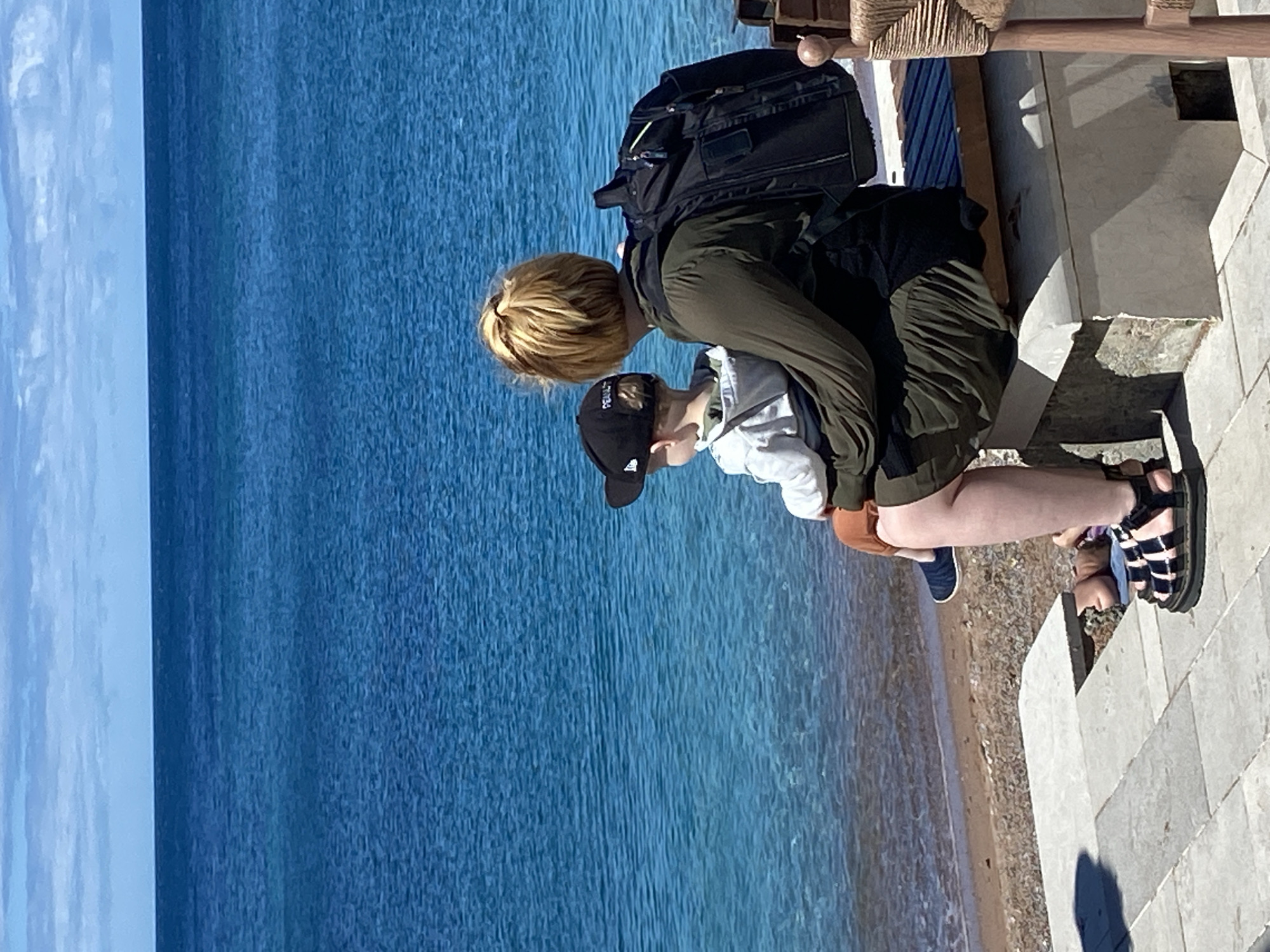 The image shows a person and a child sitting on a stone bench by the sea in Petrovac centar, Montenegro. The person, with blonde hair pulled back, wears a green top and a backpack. The child, wearing a black baseball cap, sits on the person's lap facing the water. The sea is a vibrant blue, and the sky above is partly cloudy. A beach with pebbles is visible in the foreground, leading to the water. A portion of a wooden chair is also visible on the right side of the frame.