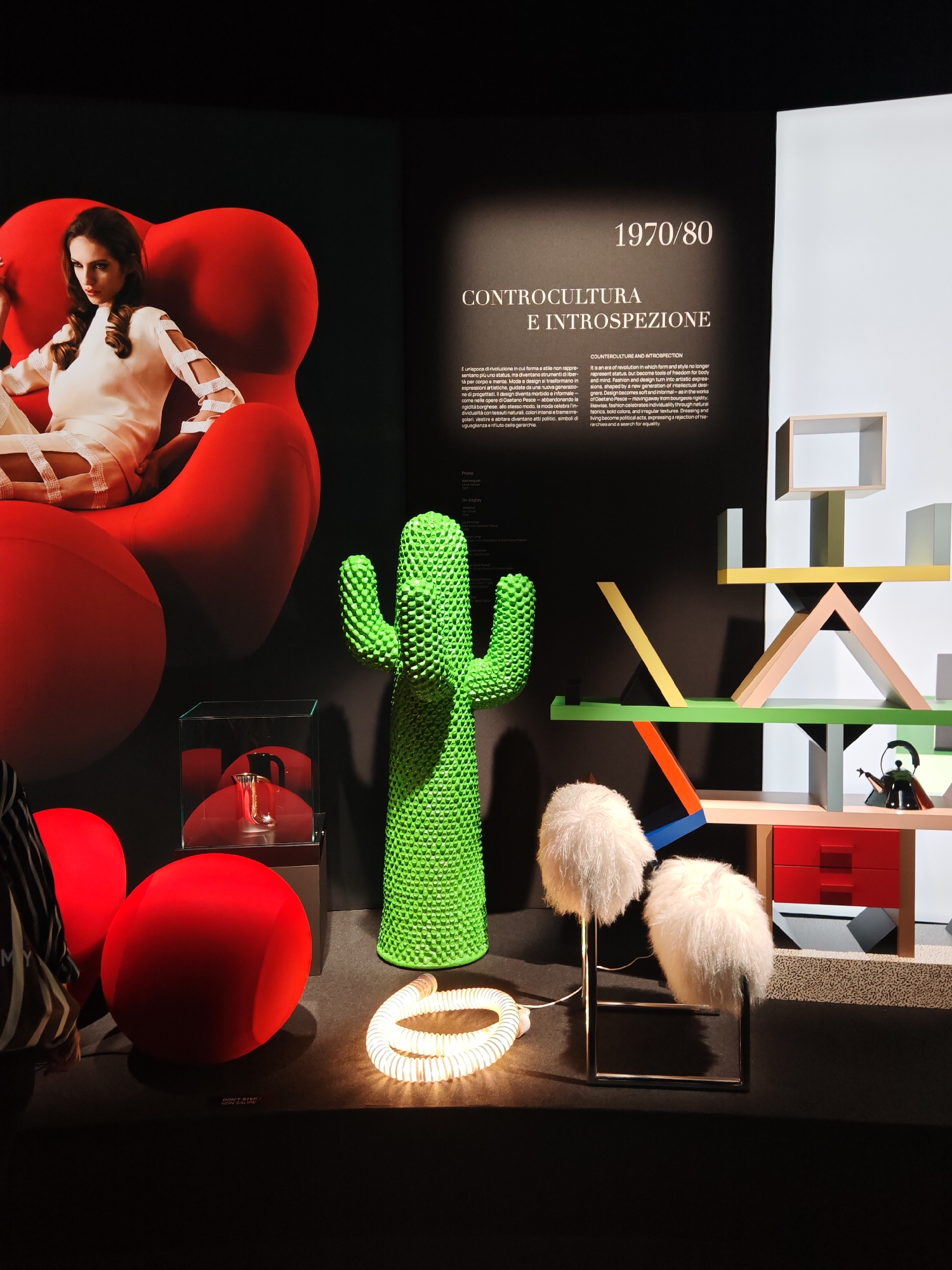 This image captures an exhibition display focused on design from the 1970s and 1980s, titled "CONTROCULTURA E INTROSPEZIONE" (Counterculture and Introspection). The scene is set in a dimly lit indoor space, likely a museum or gallery, with spotlights illuminating the exhibits.
On the left, a large poster features a woman with long, dark hair, dressed in a cream-colored outfit with cut-out sleeves, sitting poised in a striking red, organic-shaped armchair, possibly the "Up 5" by Gaetano Pesce. Below this, two smaller, round red seats or poufs are arranged on the floor.
The center of the display showcases a tall, textured, bright lime-green cactus-shaped object, known as the "Cactus coat stand" by Gufram. Beside it, a clear glass display box holds a shiny, reflective silver object. On the floor in front, a luminous, coiled white tube light emits a soft glow. Further right, two whimsical lamps with white, shaggy, furry shades and minimalist metal bases are displayed.
To the far right, a colorful, multi-level modular shelving unit, characteristic of the Memphis Group style, is prominent. It features an eclectic mix of geometric shapes and bold colors including yellow, light green, orange, blue, black, and light pink, with red drawers at the bottom. A small black kettle sits on one of its shelves.
A large informational panel in the background provides context for the era and theme in both Italian and English. The text describes the 1970s and 80s as a revolutionary period where design and fashion transformed into artistic expressions, tools for freedom, and political statements, rejecting bourgeois rigidity in favor of individuality and equality. A "DON'T STEP / NON CALPESTARE" warning is visible on the floor. A visitor, partially visible on the bottom left, adds to the sense of an active exhibition space.