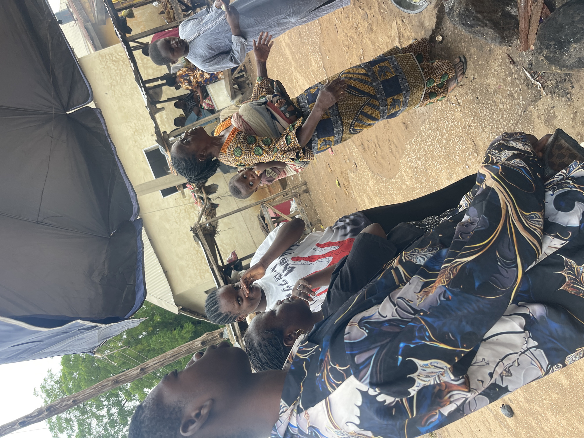 The image shows a group of people in what appears to be an outdoor setting in Chikaji, Nigeria. A woman in a patterned dress holds a child. Nearby, a group of three individuals is present; one is partially visible in the foreground wearing a patterned shirt. An umbrella provides shade in the lower left. The ground appears to be dirt or a rough, unpaved surface. In the background, there are buildings with corrugated metal roofs and what appears to be wooden scaffolding or supports. Several other individuals are further back, appearing to be engaged in conversation or observation. A man is wearing a light-colored, long-sleeved shirt.