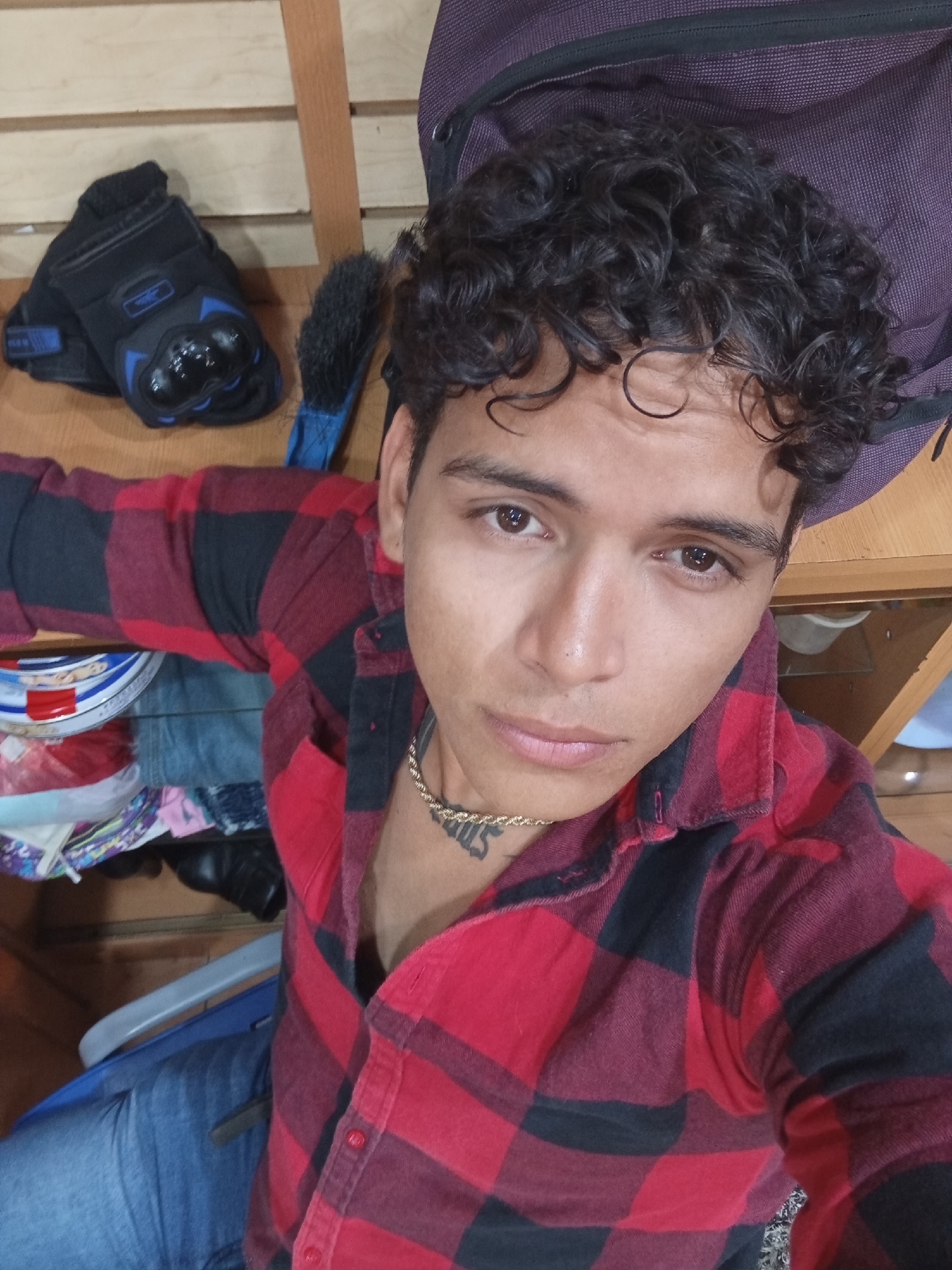 A young man, likely in his late teens or early twenties, is captured in a close-up, slightly overhead shot, appearing to be a selfie. He has dark, curly hair and brown eyes, looking directly at the camera with a neutral expression. He is wearing a red and black plaid flannel shirt, with the top few buttons open, revealing a gold rope chain necklace. On his neck/upper chest, there is a visible tattoo, which appears to spell out "SUS" or a similar word, though it is partially obscured. He is also wearing blue jeans, visible towards the bottom of the frame.
The setting appears to be indoors, possibly a room with wooden shelves or furniture in the background. To his upper left, a pair of black motorcycle gloves with blue protective plastic elements is placed on a shelf. Next to the gloves, a blue brush with black bristles is visible. Above his right shoulder, part of a dark purple backpack or bag rests on a wooden surface. Below him, on a lower shelf, various items are stacked, including what looks like clothing and a round, striped object. A portion of a blue and grey chair is also visible on the left side of the lower frame. The lighting suggests an indoor environment. The scene depicts a casual moment in Juan Griego, Venezuela.