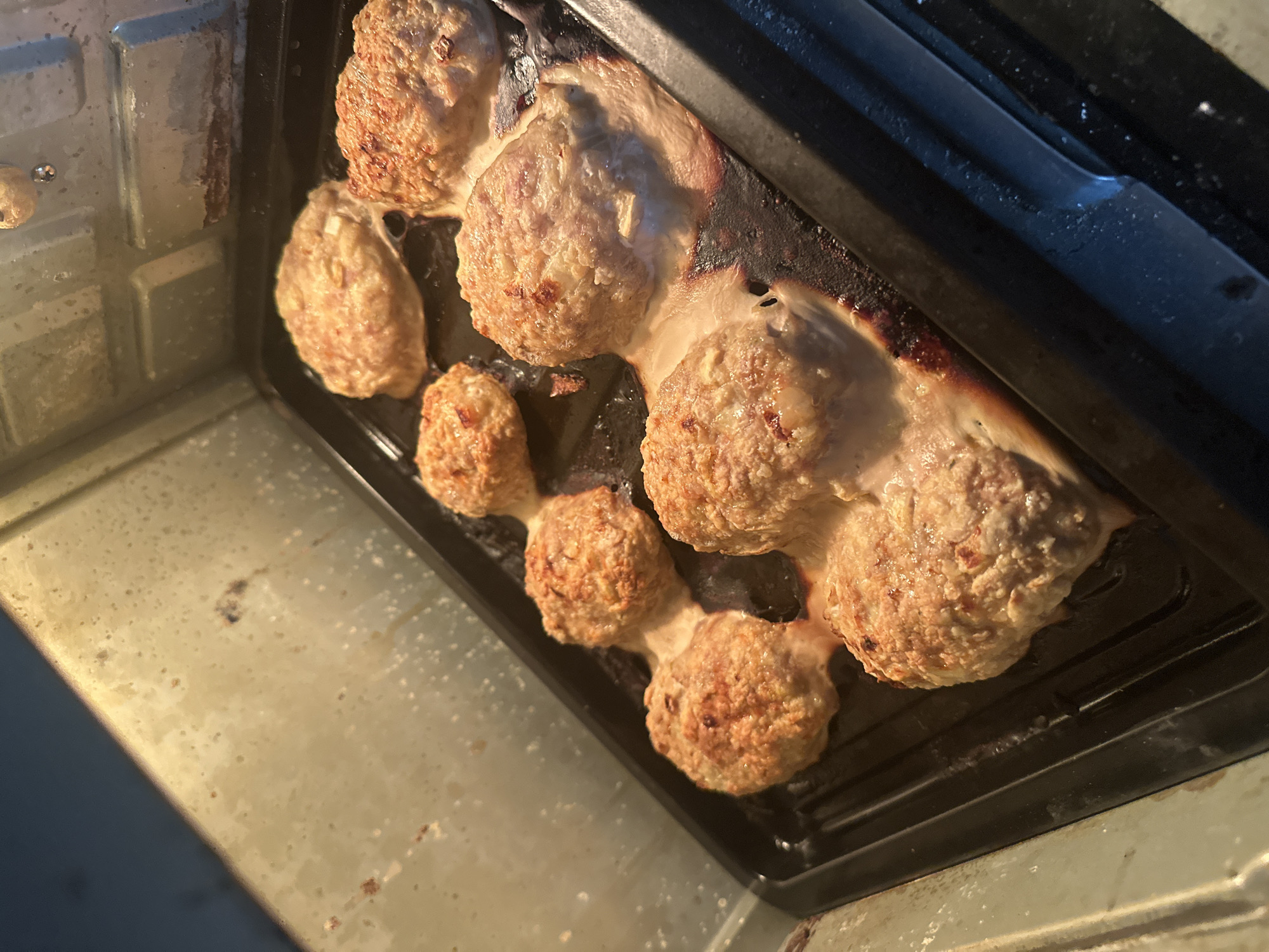A black baking pan filled with cooked meatballs sits inside an oven. A greasy, clear substance surrounds the meatballs. The inside of the oven is a pale, metallic color, suggesting it is in need of cleaning. No specific location in Bac Nha Trang Ward, Vietnam, can be determined from the image.