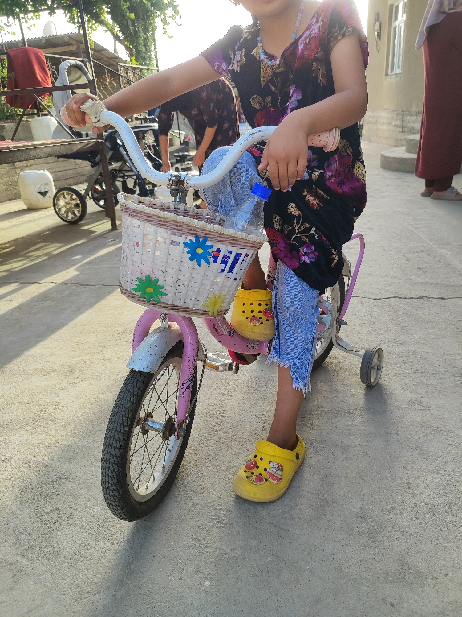 A child, partially visible, is standing next to a pink child's bicycle with training wheels. The bicycle has a white woven basket attached to the handlebars, decorated with colorful artificial flowers. The child is wearing a dark floral print dress and light blue jeans that are ripped at the hem. Their feet are clad in yellow clogs adorned with fruit and cupcake designs. The scene takes place outdoors on a paved surface that appears to be cracked in places. In the background, there is a building with a white-framed window and a glimpse of a person in a maroon outfit. The environment suggests a residential area in Sultonobod, Tajikistan, possibly a courtyard or alleyway, with some foliage and structures visible. The lighting indicates it is daytime, with sunlight casting shadows on the ground.