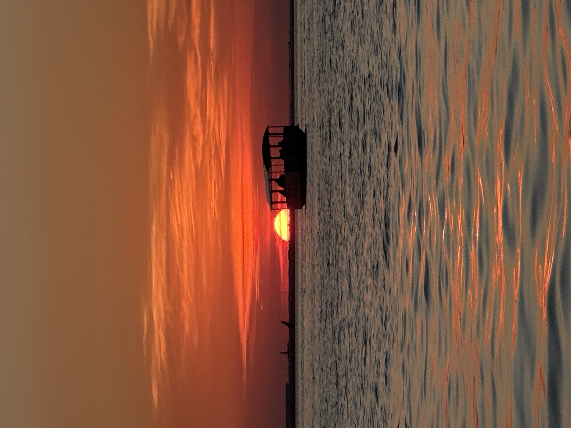 The image depicts a sunset over a body of water. The sky is a vibrant gradient of orange and red, with visible horizontal cloud formations. A small boat with a covered top, silhouetted against the sky, is positioned in the middle of the water. Several figures are visible as silhouettes within the boat. In the distance, a dark horizontal line represents the shoreline, with a faint tower or structure visible on the left. The water reflects the colors of the sunset, creating shimmering orange streaks across its surface. The scene is peaceful and serene. There is no visual information in the image that allows for the identification of the specific location in Kokshetau, Kazakhstan.