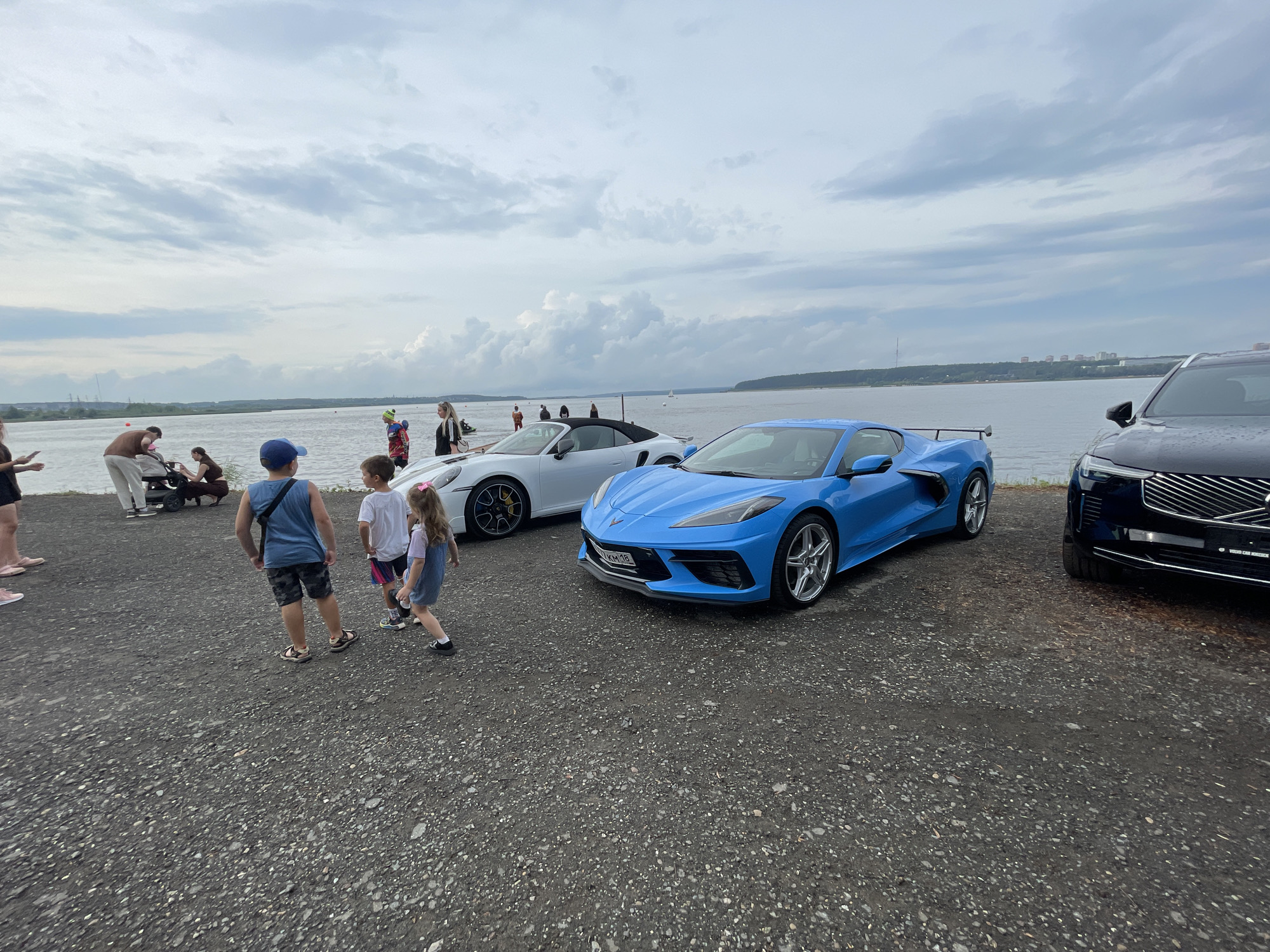 The image depicts an outdoor scene with several vehicles and people by a body of water, possibly a river or lake, under a cloudy sky. In the foreground, on a gravel surface, are two sports cars: a bright blue Chevrolet Corvette with a visible license plate and a white convertible, which appears to be a Porsche. To the right, the front of a dark SUV or crossover vehicle is visible, also on the gravel.
Several people are present. In the middle ground, three children are walking. One boy in a blue tank top and patterned shorts is walking away from the camera. A second boy in a white t-shirt and shorts is walking towards the right. A young girl with a pink bow in her hair, wearing a denim dress, is walking towards the left. Further back, near the white convertible, a group of people are gathered, including individuals pushing a stroller and standing around. Some figures are seen standing near the water's edge in the background. The background shows a wide expanse of water, a distant shoreline with trees and buildings, and a hazy cityscape. The overall impression is a casual gathering or event, potentially a car show, by the water. The image is taken from a ground-level perspective.