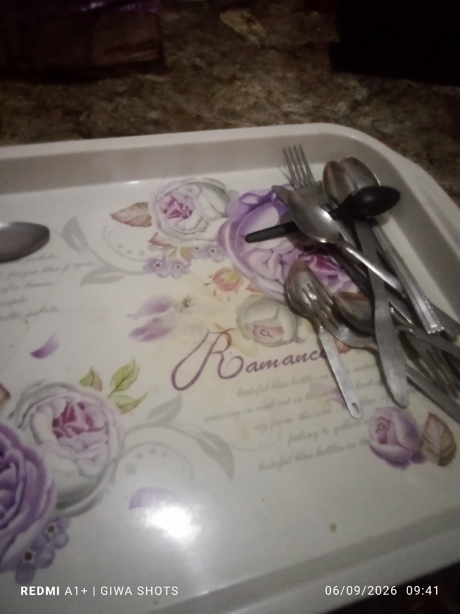 A zoomed-in, slightly out-of-focus, eye-level shot captures a white plastic tray adorned with a floral pattern, primarily featuring purple and pink roses. Several metallic utensils, including spoons and a fork, are arranged haphazardly on the tray. The background is dark and indistinct, suggesting an indoor setting, possibly a kitchen or dining area. The bottom left corner displays the text "REDMI A1+ | GIWA SHOTS," and the bottom right corner shows the date "06/09/2026" and time "09:41." No specific landmarks in Jalingo, Nigeria are visible.