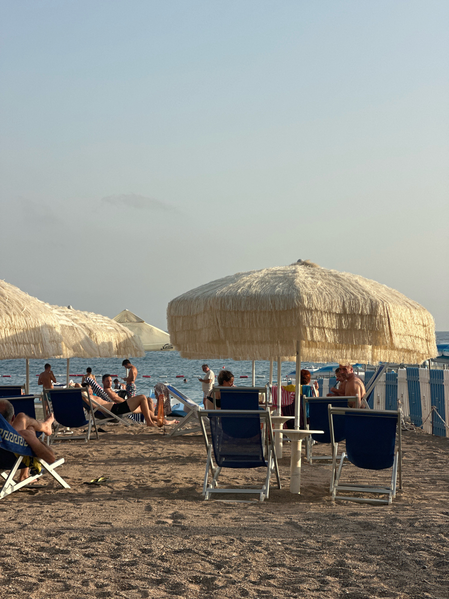 The image depicts a beach scene in Boreti, Montenegro. Several people are visible, some lounging in chairs under large, thatched umbrellas. The sand is coarse, and lounge chairs with blue fabric are set up on the beach. In the background, the sea is calm, with some people visible in the water. A striped barrier with blue and white fencing runs along the edge of the beach, separating it from the water. The sky is a pale blue with some wispy clouds. One person in the foreground is reclining in a lounge chair, looking at their phone. Another person, a man, is sitting up in a chair, also looking at their phone. A group of people are gathered under a large umbrella, some standing and some seated. The overall impression is one of a relaxed beach day.