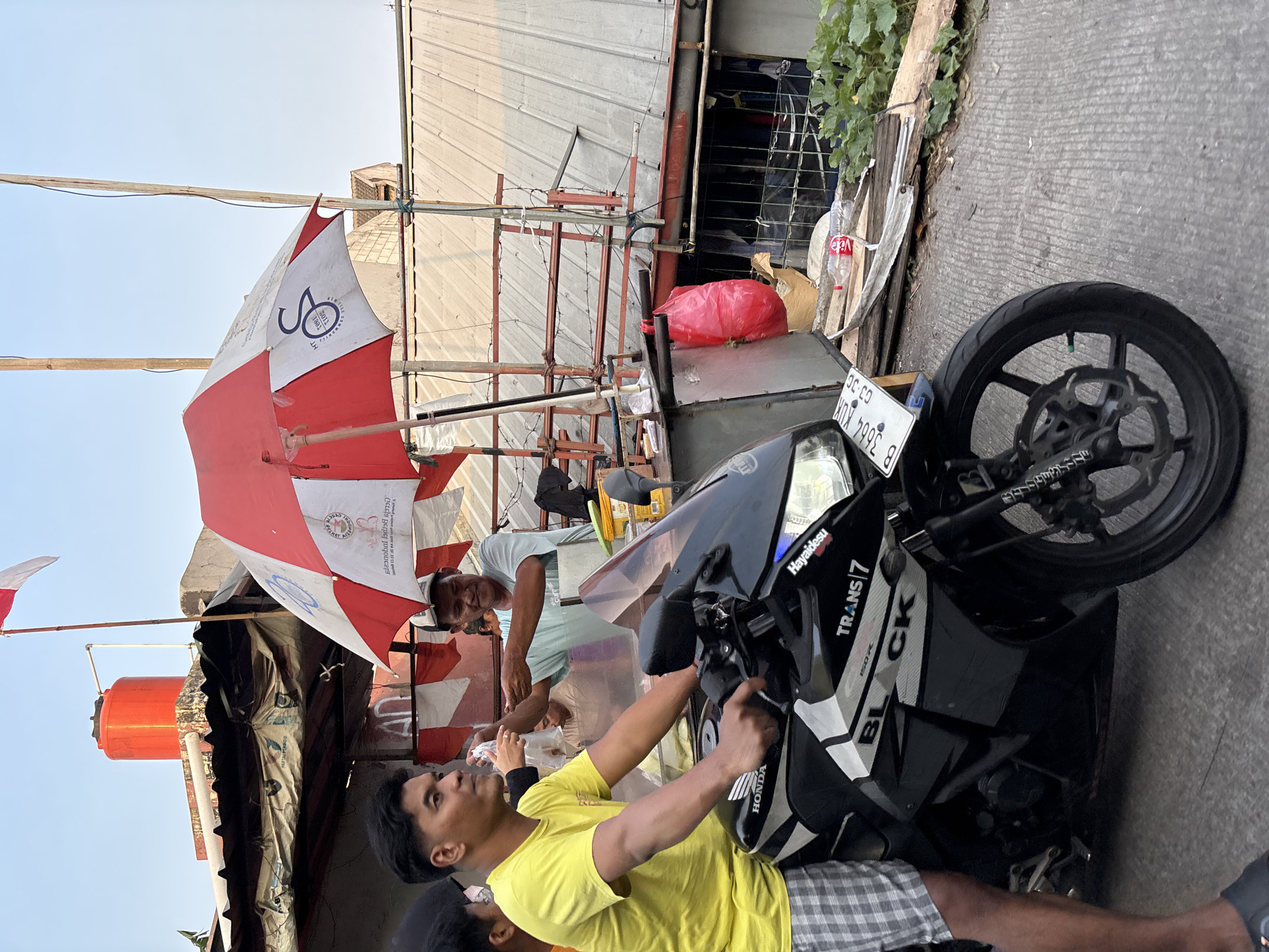 In Bekasi, Indonesia, a man wearing a yellow t-shirt and checkered shorts rides a black Honda motorcycle. He is looking to his right, where another man, wearing a light blue t-shirt and a cap, is handing him something from behind a red and white umbrella. The umbrella is supported by poles and attached to a structure with a corrugated metal roof. Behind them, an orange water tank is visible on the roof of a building, and a red and white Indonesian flag is flying from a pole. The scene appears to be on a street with a concrete surface, and there are some plants and debris on the side of the road. The motorcycle has a license plate that reads "B 3664 KUX".