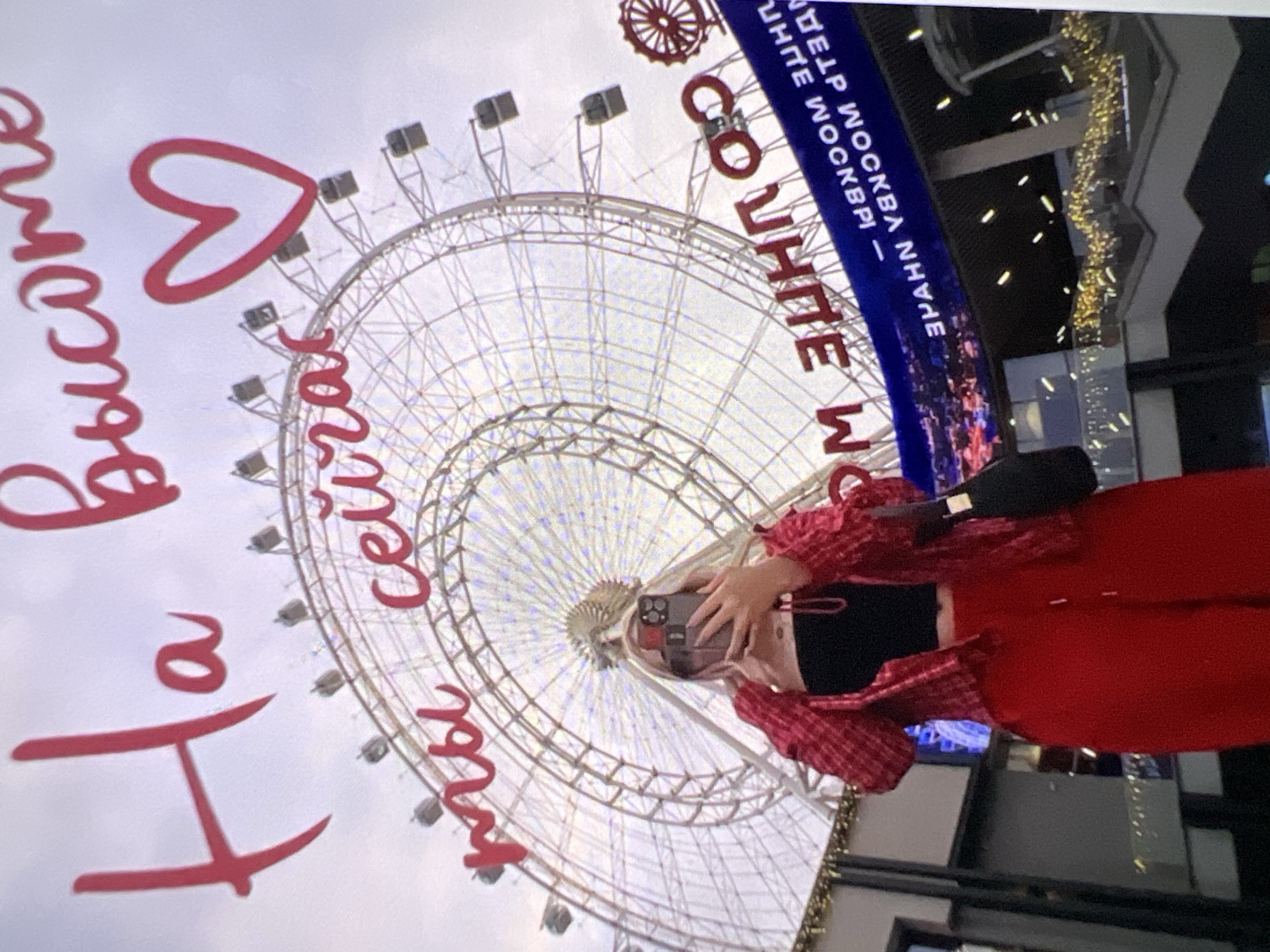 A person is seen taking a photo or selfie in front of a large Ferris wheel under an overcast sky. The person, likely female, wears a black crop top, a red and white plaid long-sleeved shirt, and red pants, with a small black bag slung over their shoulder. They hold up a smartphone with a multi-lens camera to capture the scene.
The Ferris wheel, a prominent feature of the background, is a massive structure with white or light-colored spokes and gondolas. To the right, a building or sign with blue and white lettering is visible, adorned with warm-toned lights. The sky is a uniform light gray, suggesting a cloudy day.
Visible text includes:
* Red cursive text superimposed over the image, which reads "На высоте" (Na vysote), meaning "At a height," and partially visible below it, "ты сейчас" (ty seychas), meaning "you are now," forming "You are now at a height."
* White block letters on the blue background of a sign, which appears to say "ВДНХ МОСКВА НА ВЫСОТЕ МОСКВА" (VDNKh Moskva Na Vysote Moskva), translating to "VDNKh Moscow at the height of Moscow." Below this, "SO WHITE MW" or a similar phrase in English is partially visible.
The scene suggests a visit to a prominent landmark or amusement area, likely VDNKh in Moscow, given the Russian text and the presence of a large Ferris wheel.