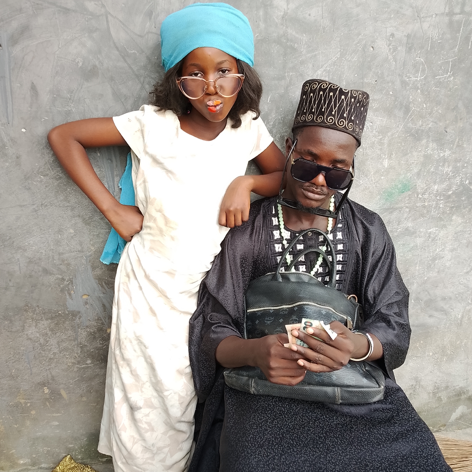 In Ado, Nigeria, two individuals are posed against a weathered, grey concrete wall. On the left, a young girl, wearing a bright blue headscarf and white dress, leans against the wall, her arm resting on the shoulder of the person beside her. She sports large, rose-gold framed glasses and has a colorful item, possibly candy, at her lips. To the right, a man, dressed in a black traditional outfit with intricate gold patterns and a matching black cap adorned with similar gold embroidery, is seated. He wears dark sunglasses, and around his neck are light green beaded necklaces. He is holding several banknotes, seemingly counting or displaying them, with a black leather bag, featuring a prominent MCM logo, resting in his lap. The environment appears to be outdoors, with a portion of a broom visible at the bottom right.