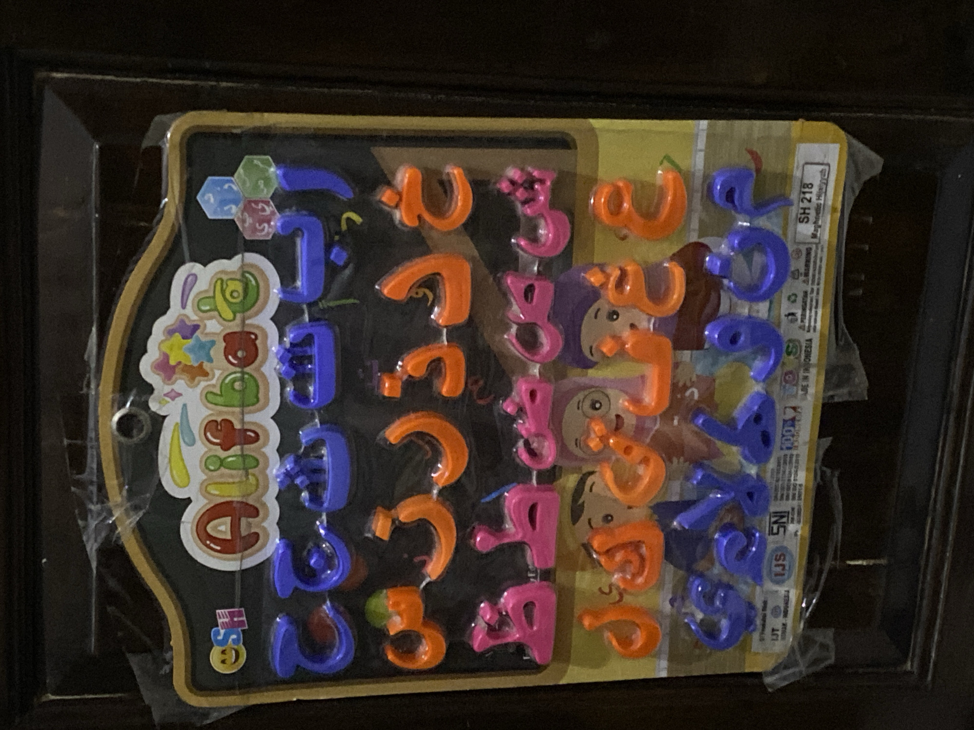 A magnetic Arabic alphabet learning toy is displayed in its original plastic packaging. The toy features colorful, three-dimensional magnetic letters, predominantly in blue, orange, and pink, arranged in rows on a black background designed to resemble a chalkboard. The packaging also includes a colorful title, "Alifba," and several cartoon illustrations of children. The product information at the bottom indicates it is a "Magnetic Hijaiyah" toy, model "SH 218," manufactured in Indonesia, and is SNI certified. The toy is positioned against a dark, textured surface, likely a wooden door or furniture. The scene appears to be an indoor setting, possibly a home or a shop, in Pekalongan, Indonesia, suggested by the SNI certification and "Made in Indonesia" markings. There are no people actively interacting with the toy.