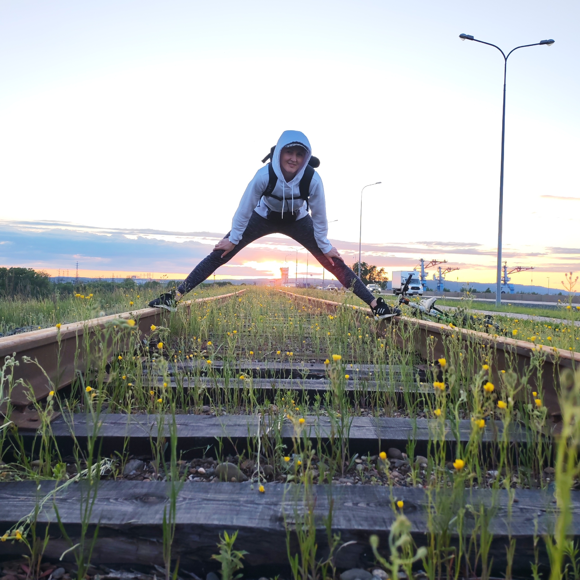 An individual, likely a young adult, is captured in a playful pose on overgrown railway tracks during either sunset or sunrise in Chaikovsky, Russia. The person, wearing a light blue hoodie, dark patterned leggings, and black athletic shoes, stands with a wide stance, one foot on each rusted rail, and hands resting on their knees. A black backpack is visible on their back, and they look towards the camera with a slight smile. The low-hanging sun, a bright orange-yellow glow, is centered on the horizon behind the person, casting a warm light over the scene.
The foreground is dominated by the disused railway tracks, with wooden sleepers largely covered by green grass, weeds, and numerous small yellow wildflowers. A bicycle lies on the ground next to the right-hand track. In the mid-ground, a road or highway runs parallel to the tracks, with a white truck and other vehicles visible. Tall streetlights line the road. The background features industrial structures, possibly cranes or transmission towers, silhouetted against a sky painted with warm orange, pink, and light blue hues, indicative of dusk or dawn. The overall setting is an outdoor, somewhat industrial landscape on the edge of a populated area, suggesting a moment of leisure or adventure. No visible text is present in the image.