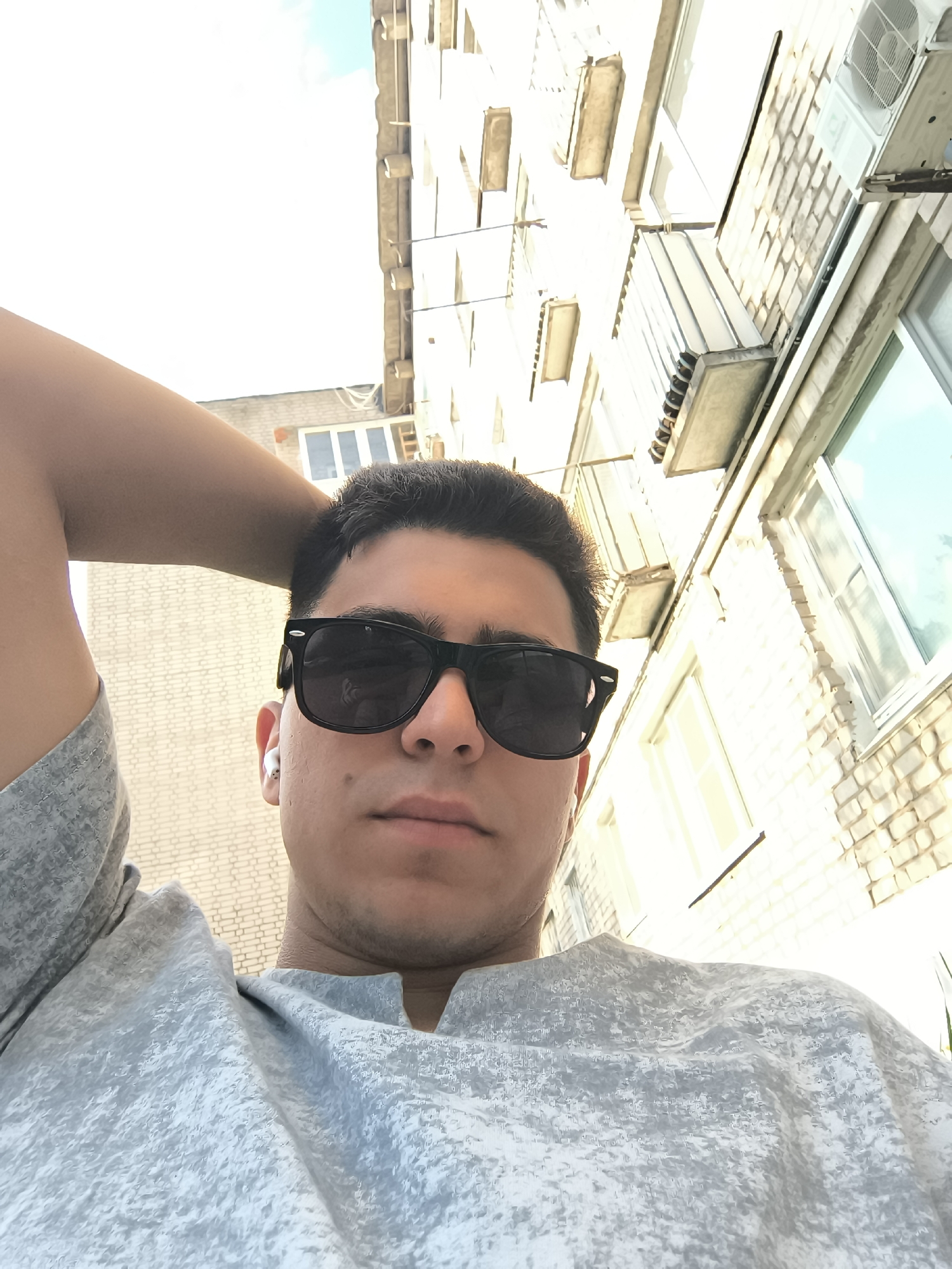 A selfie shows a man with dark hair and sunglasses, his left arm raised behind his head. He is wearing a light gray shirt with a V-neck. A white wireless earbud is visible in his left ear. In the background, a multi-story brick building can be seen, featuring multiple windows and air conditioning units. The perspective is from below, looking upwards towards the man and the building. The location is likely an urban area, though specific street details are unavailable. The lighting suggests a daytime shot.