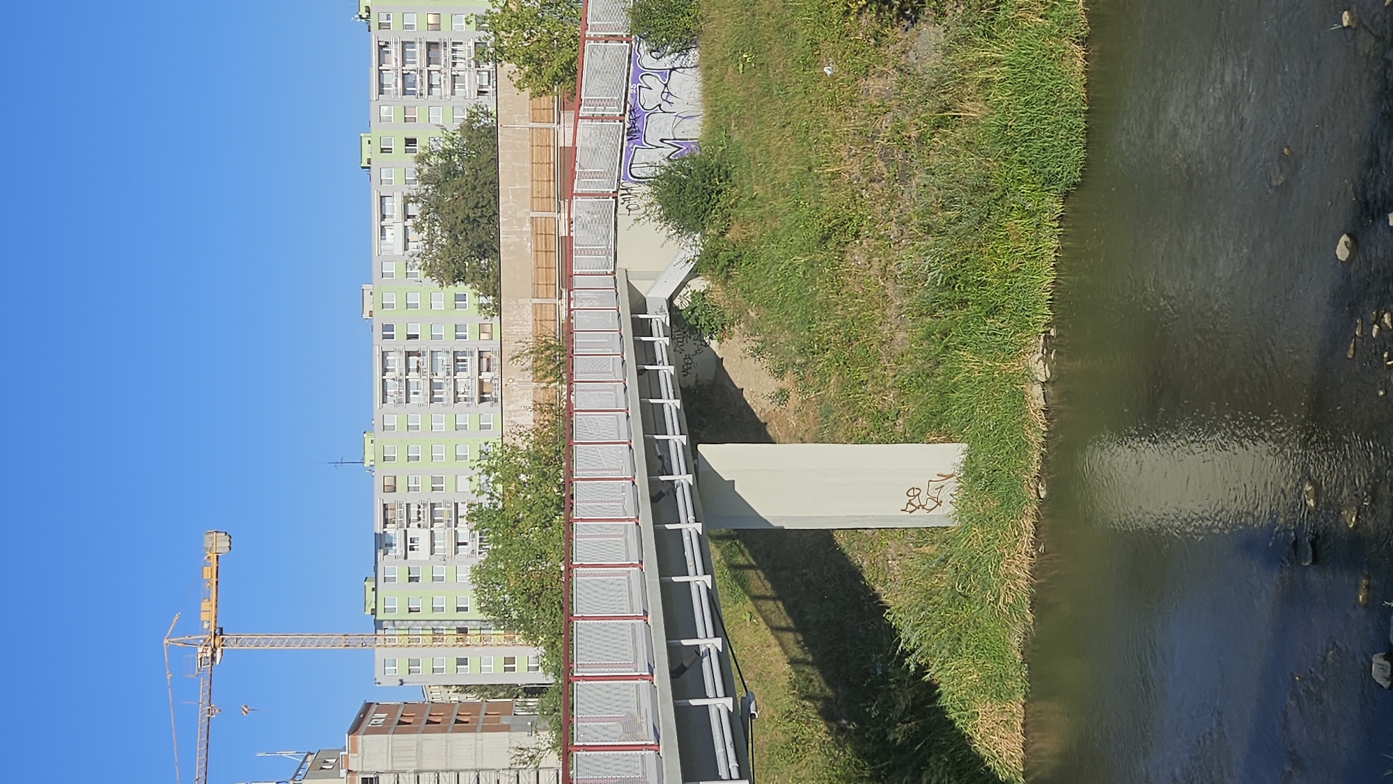 This image shows a pedestrian bridge over a river in Prešov, Slovakia. The bridge has a grey deck with white mesh railing and red top and bottom rails. The bridge is supported by a concrete pillar. There is grass and vegetation on the riverbanks, with some graffiti on the concrete pillar and the embankment. In the background, there is a light green apartment building with many windows and balconies, and a construction crane is visible to the left of the apartment building. The sky is clear and blue.