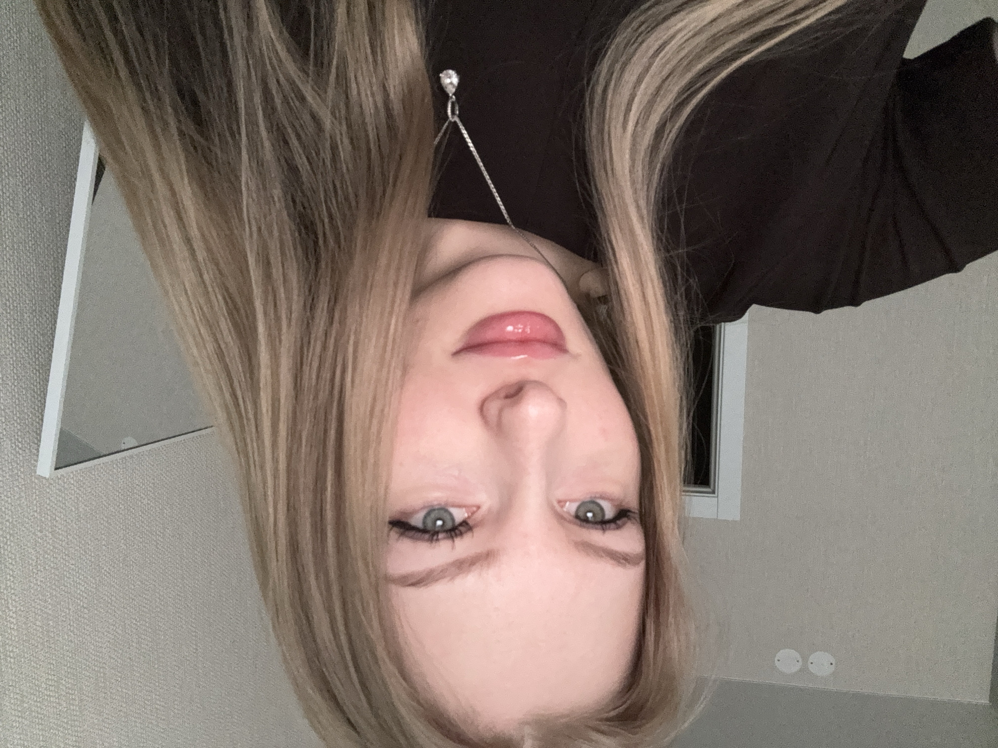 A young woman with long, blonde hair is shown in a close-up, upside-down portrait. She has fair skin, light-colored eyes, and pink, glossy lips. She is wearing a dark, long-sleeved shirt. A silver necklace with a teardrop-shaped pendant is visible. The background consists of a wall with a light-colored textured pattern. A white-framed mirror is visible in the upper left corner, and a window with a white frame is in the upper right. Two white circular objects are visible on the wall in the lower right. The location is in Perm, Russia.