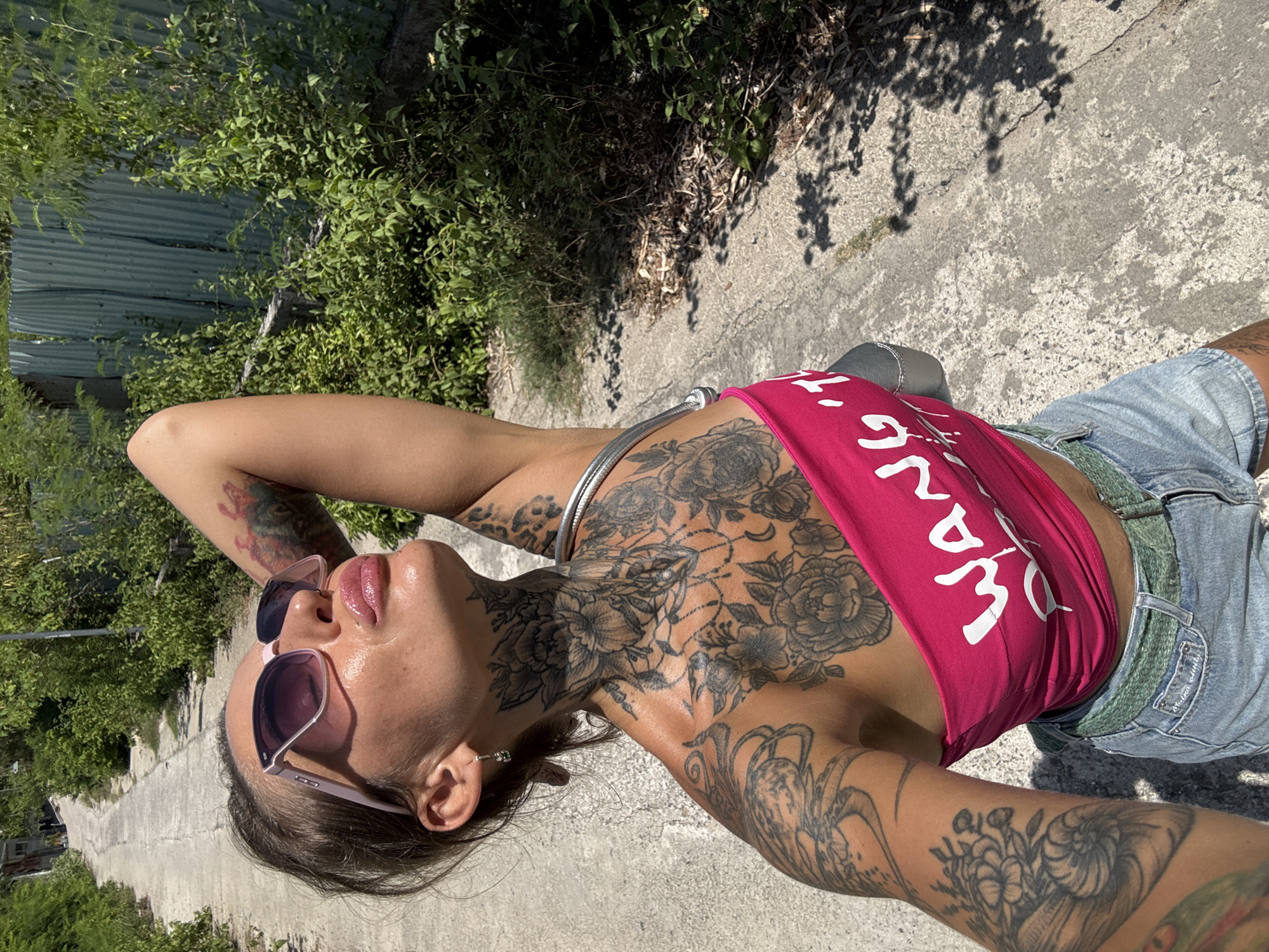In a sun-drenched alleyway in Bac Nha Trang Ward, Vietnam, a woman with extensive tattoos poses for a selfie. She wears a bright pink tube top with white lettering, denim shorts, and sunglasses. Lush green foliage flanks the concrete path, and a corrugated metal fence is visible in the background. The woman's arm is raised behind her head, and her body is angled towards the camera.