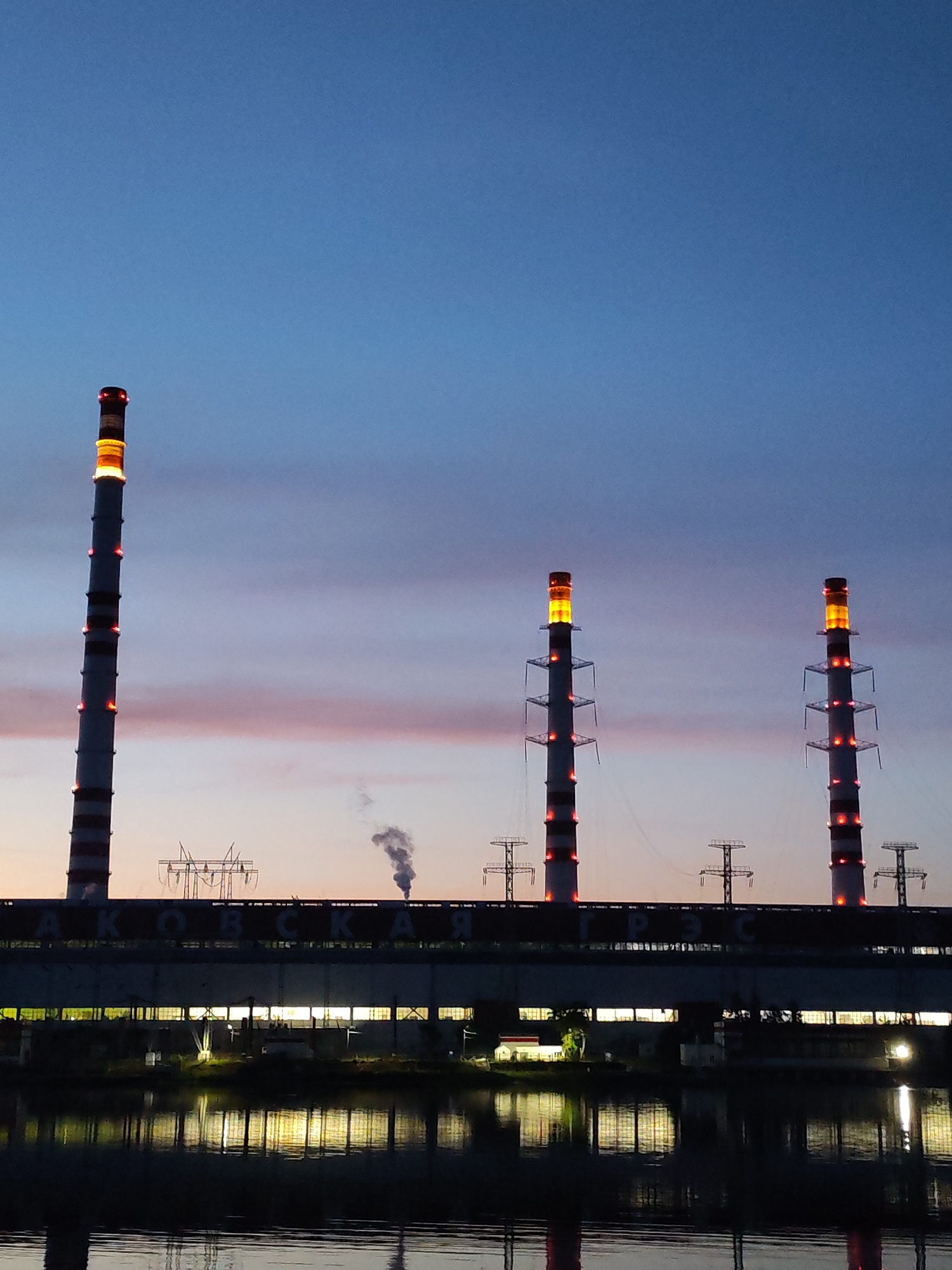 The image depicts a power plant at dusk. Three tall smokestacks dominate the scene, their upper sections illuminated with red and orange lights. The smokestacks are part of a long, industrial building that stretches across the middle of the frame. The building's windows are lit from within, casting a warm glow onto the water below. A river or large body of water runs in the foreground, reflecting the lights of the building and the smokestacks. Smoke plumes rise from one of the smokestacks, indicating ongoing operations. The sky above is a deep blue, transitioning to lighter shades of purple and pink near the horizon, characteristic of twilight. The overall impression is one of industrial activity set against a serene evening sky.
