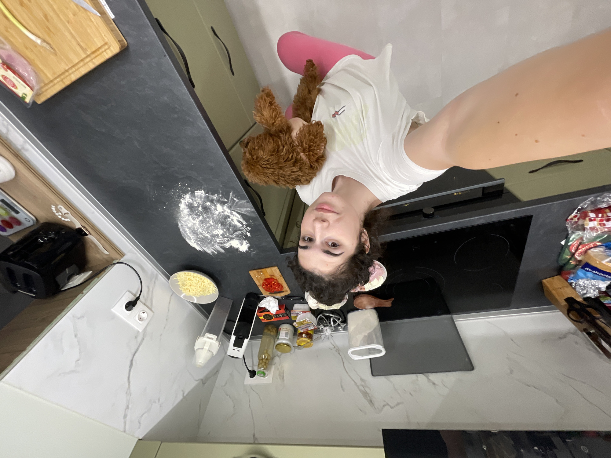 A woman is holding a small brown poodle in her left arm, standing in a kitchen. She is wearing pink pants and a white t-shirt. The kitchen has white marble countertops with a black stovetop and black countertop to the left. On the black countertop, there is flour, cheese, and chopped red peppers. There are also canisters and bottles on the counter. In the background, there is a cutting board with food and kitchen cabinets. The location is likely an apartment in Sofia, Bulgaria.