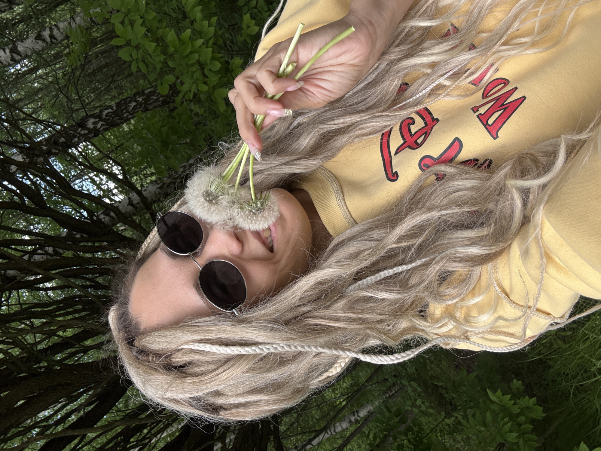A female person with dreadlocks wears sunglasses and a yellow sweatshirt. She holds a bouquet of dandelions in front of her face. The location is a park with trees. The city and state are Unknown.