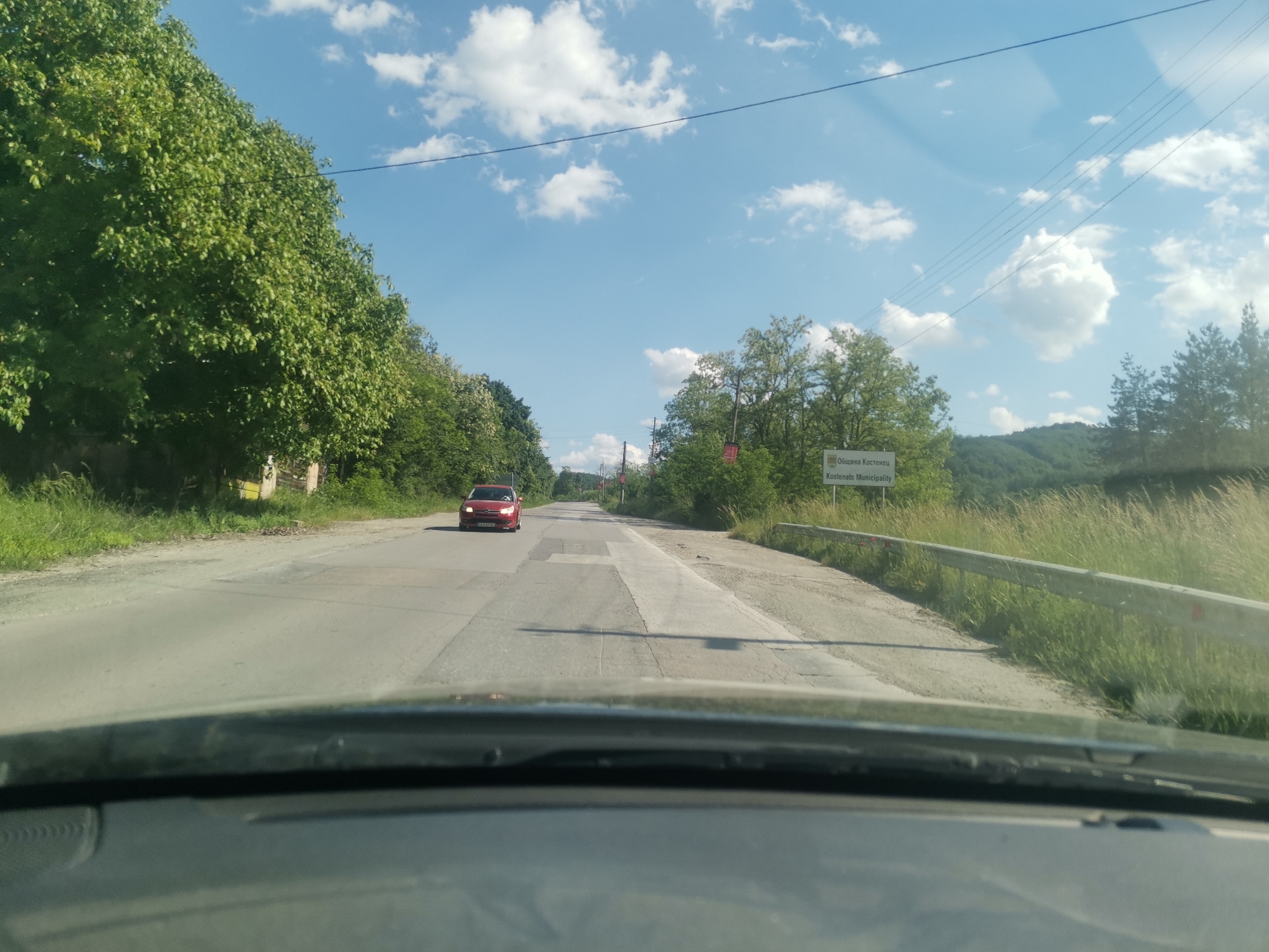 The image shows a road scene in Momin Prohod, Bulgaria. The view is from inside a car, looking out onto a paved road. A red car is approaching in the opposite lane. The roadside features green foliage, including trees and grassy areas. A sign indicates the location is the "Kostenets Municipality." The sky is blue with scattered white clouds. There are power lines overhead. The foreground shows the dashboard of the car.