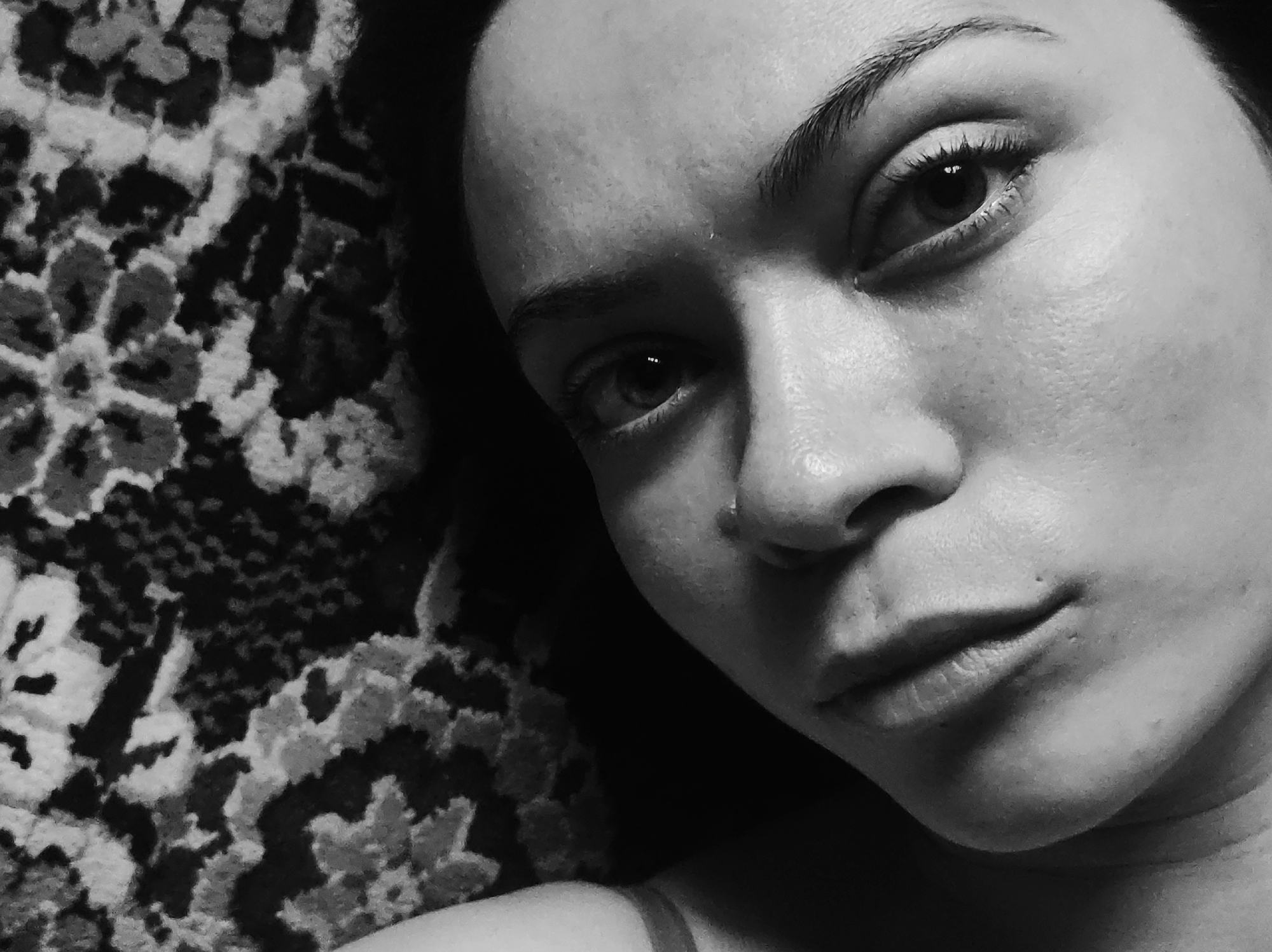 A black and white close-up of a person's face. The person is lying on a surface with a patterned texture. The person has dark eyebrows, eyes, and dark hair. They have a piercing on their nose. The location is Unknown, Unknown.