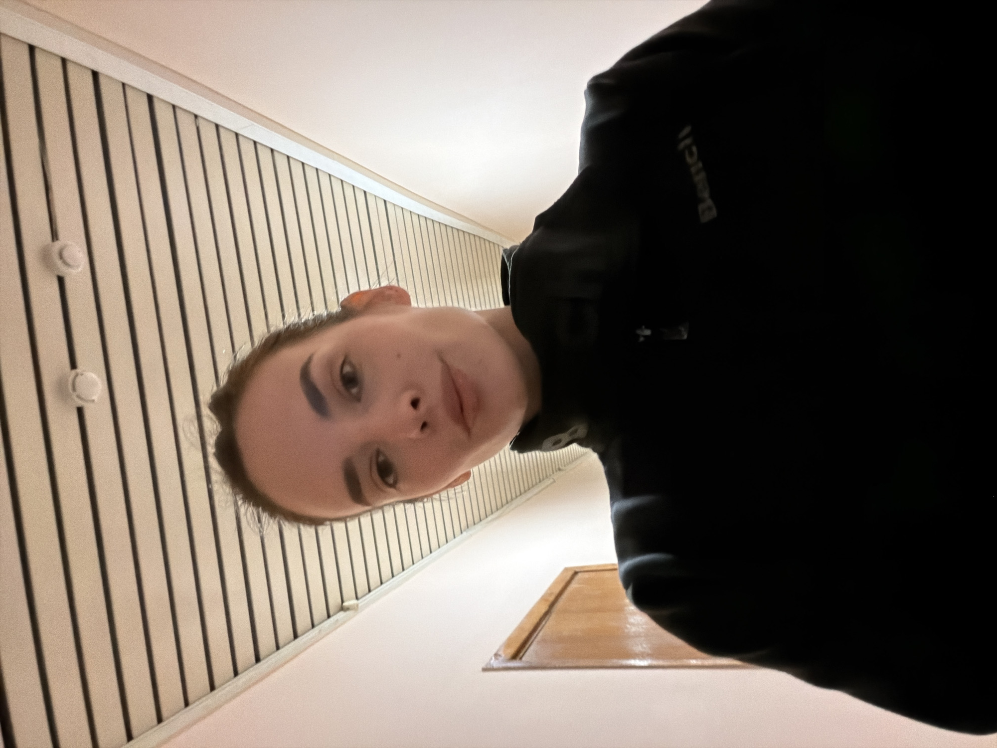 A young woman is standing in a narrow hallway, looking up at the camera. The hallway has pale pink walls and a slatted ceiling. There is a wooden door on the left side of the frame. The woman is wearing a black zip-up jacket with the word "Bench" embroidered on it in a light color. Her hair is pulled back in a bun, and she is looking directly at the camera with a neutral expression. The image appears to be a selfie, taken from a low angle. The location is Ufa, Russia.
