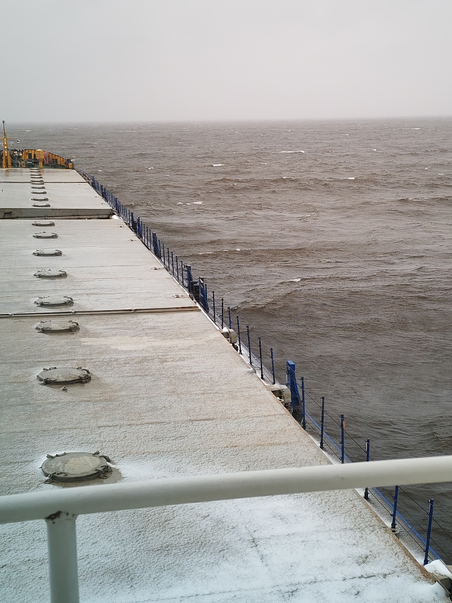 The image presents a view from the deck of a cargo ship navigating a large body of water on a cold, overcast day, likely near Shcheleiki, Russia. The ship's deck, occupying the foreground and left side of the frame, is covered in a thin layer of snow or ice. Numerous circular, bolted hatch covers are visible along the length of the deck. A blue metal railing system with vertical stanchions and horizontal wires runs along the starboard edge of the deck, bordering the water. In the immediate foreground, a white handrail is partially visible.
The water appears dark grey and turbulent, with numerous small waves and whitecaps indicating windy conditions. The horizon is indistinct, blending the water with a grey, heavily clouded, or misty sky, suggesting poor visibility. Towards the aft section of the ship on the left, some yellow equipment or superstructure elements are visible. The overall scene conveys a sense of winter travel and a harsh, cold marine environment. No people or discernible text are visible in the image.