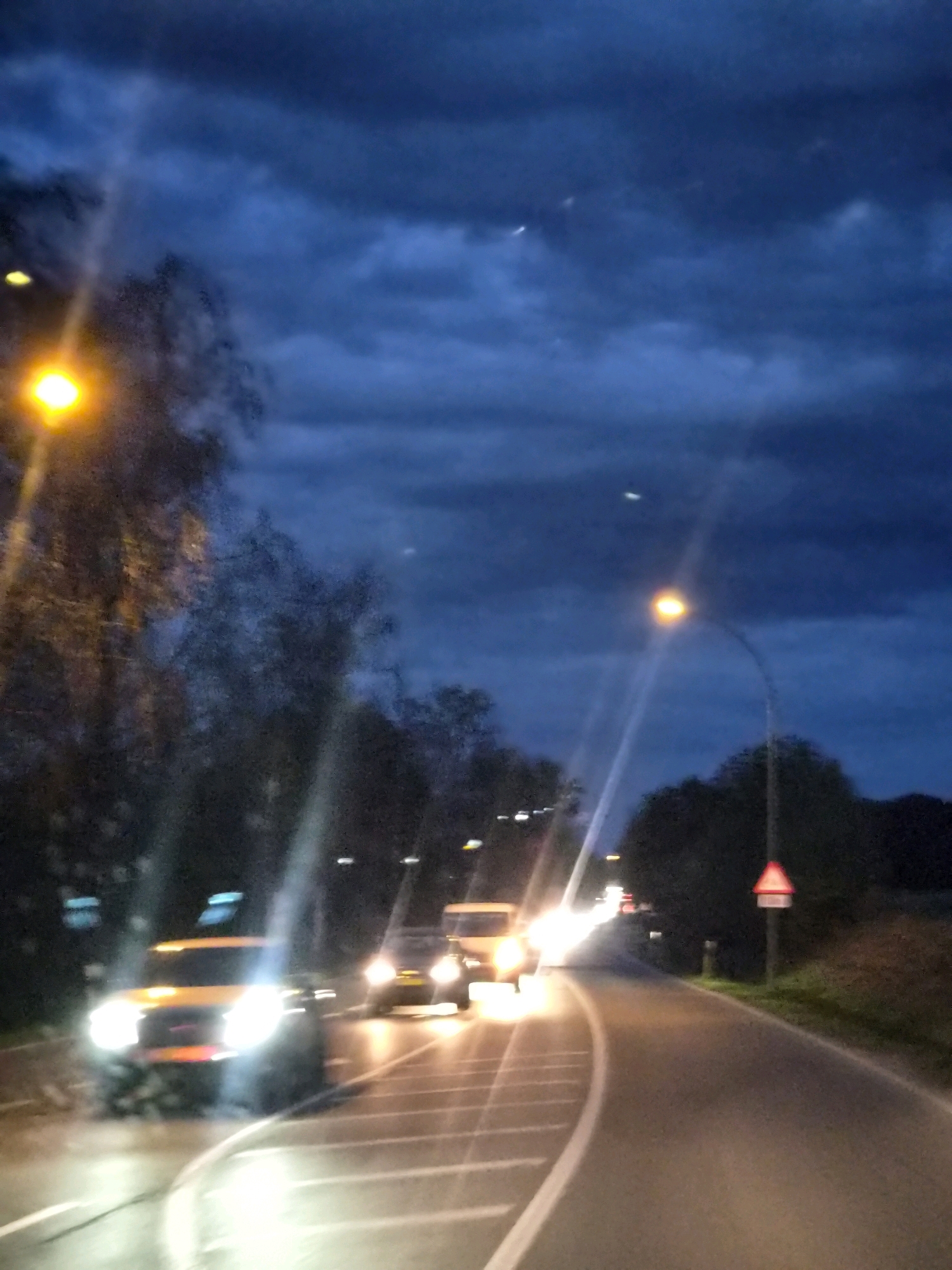 The image depicts a scene on a road in Leudelange, Luxembourg, during twilight or at night. The sky is dark blue with visible clouds. A bright street light illuminates the left side of the frame, casting light on silhouetted trees and a blurred vehicle. On the road, several vehicles are visible. A yellow SUV with its headlights on is in the foreground, moving towards the viewer. Behind it, other vehicles are seen with their lights illuminated, suggesting traffic. A traffic sign, triangular with a red border and white interior, is visible on the right side of the road. The road surface appears to be asphalt, with white lane markings. The overall impression is of a street scene with moderate vehicular traffic in low-light conditions.