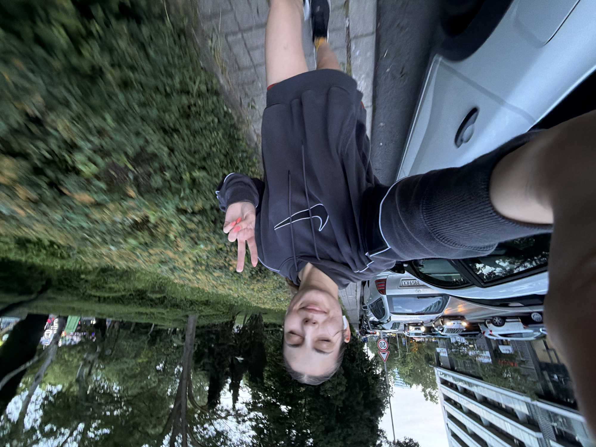 This media file captures a selfie of a young person, likely female, taken outdoors. The subject is positioned in the lower part of the frame, smiling with her eyes closed and making a peace sign with her right hand, which has orange nail polish. She is wearing a dark grey or black hoodie featuring a white Nike swoosh logo, and black shorts with a visible red waistband. White earbuds are visible in her ears.
The background reveals an urban or suburban street scene. To the subject's left, a white Mercedes-Benz car with the license plate "3959KX" is parked on the street, alongside other vehicles further down. A red circular road sign indicating a "20" speed limit is prominently visible near the street. To her right and extending upwards, lush green trees and dense foliage create a natural contrast to the built environment. In the distance, multi-story buildings can be seen under what appears to be a clear sky.
The lighting suggests it is daytime, possibly late afternoon, with soft, ambient light. The overall mood conveyed by the subject's expression and gesture is relaxed and happy. Given the provided context, the scene is located in Burgas, Bulgaria.