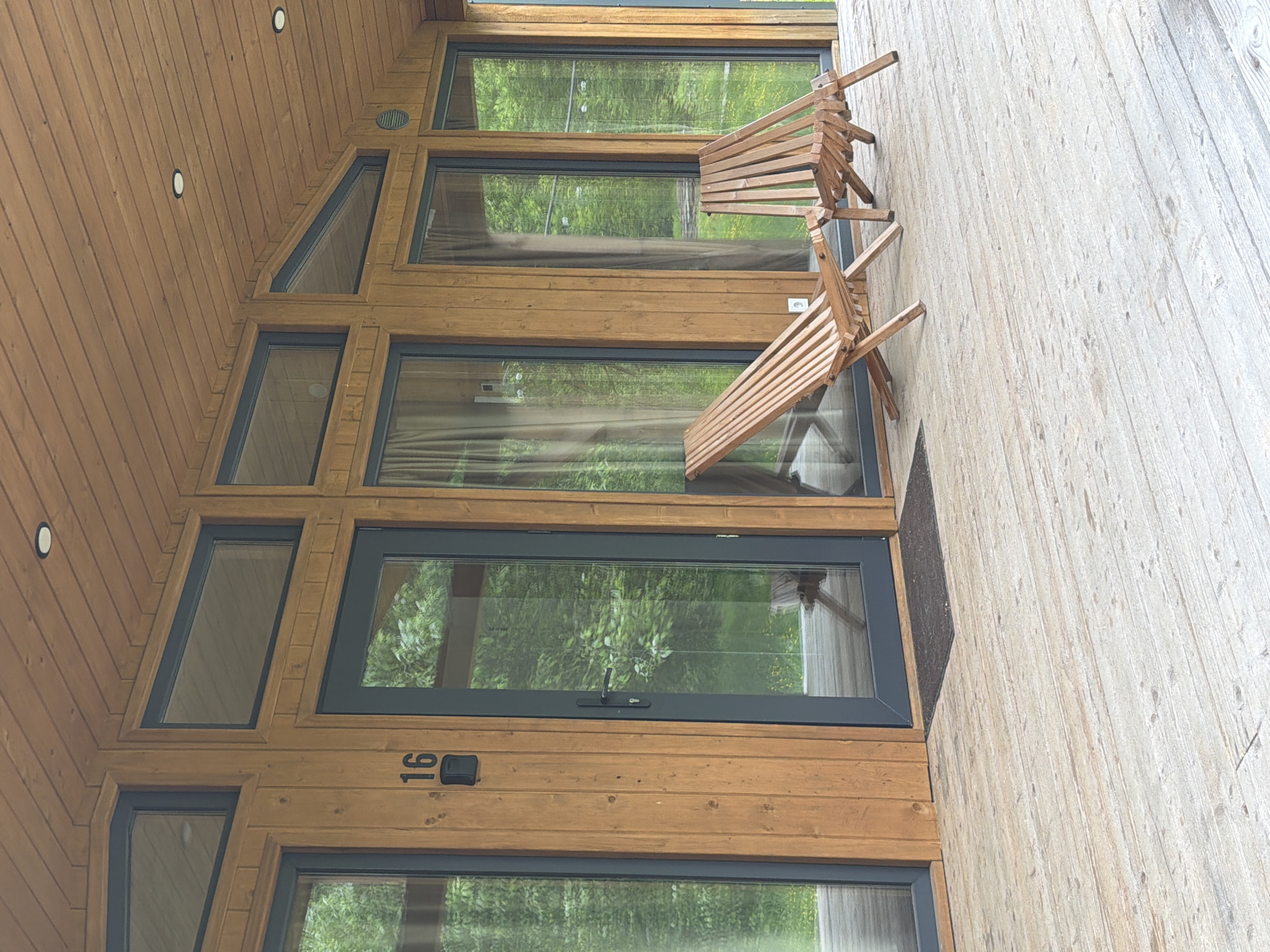 The image displays the exterior of a modern wooden cabin or building, likely located in a natural, wooded environment within Vozhoiskii, Russia. The structure's facade is clad in warm-toned horizontal wooden planks. It features a complex arrangement of numerous windows with dark gray frames, including rectangular and trapezoidal shapes, particularly in the upper section. These windows reflect lush green foliage, indicating a dense outdoor setting. Light-colored curtains are visible behind some of the panes.
On the adjacent light-colored wooden deck, two wooden slatted deck chairs are present; one stands upright, while the other is tilted or partially folded. A glass-paneled door, also with a dark gray frame and a handle, is positioned near the bottom of the structure, with a dark doormat placed in front of it. The number "16" is prominently affixed to the wooden wall above this door. Two circular recessed lights are visible on the wooden ceiling or soffit above the window array. No people are visible, and no specific activity is taking place. The overall lighting suggests it is daytime, possibly under overcast skies, providing soft, diffused illumination.