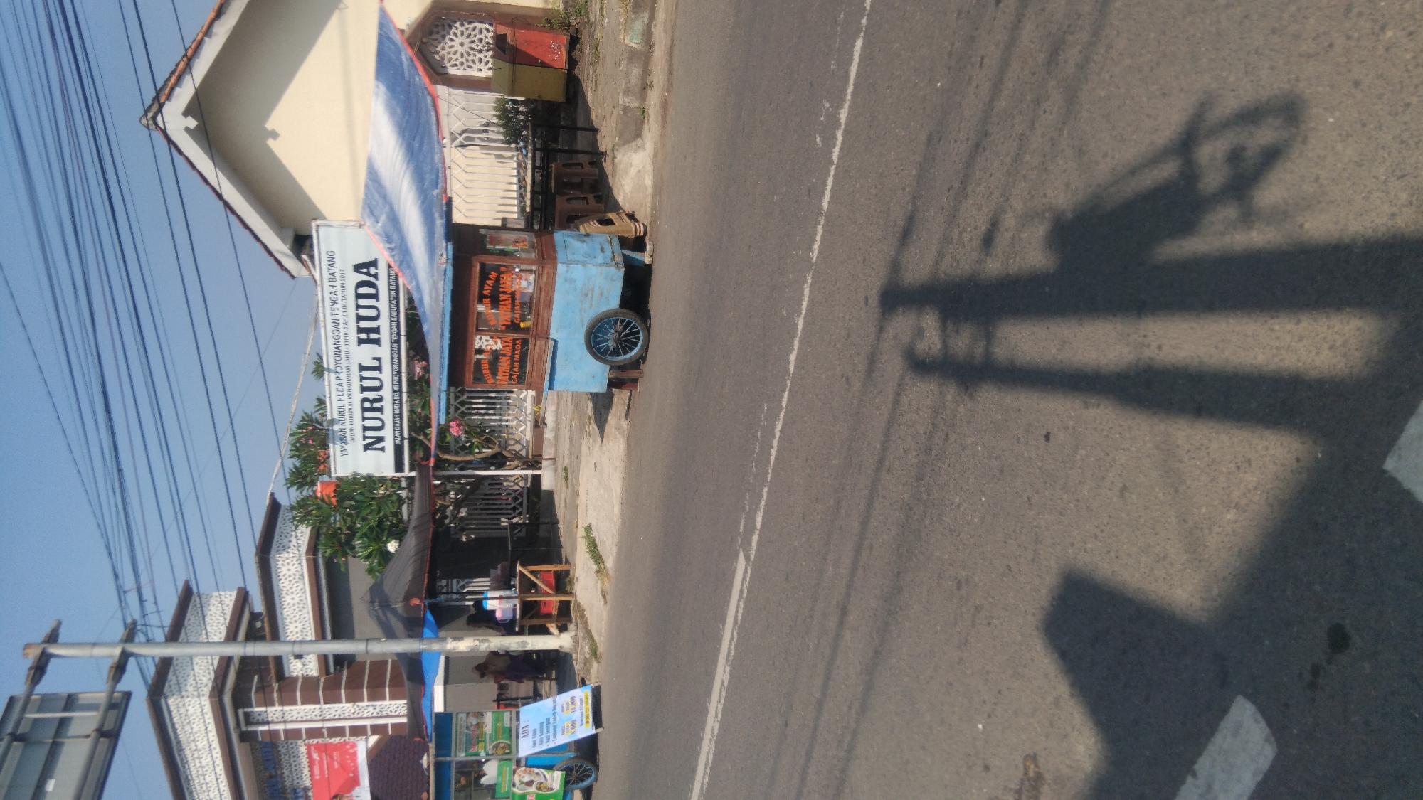A street scene in Proyonanggan Tengah, Indonesia, on a clear day. The image captures a roadside, with a paved road in the foreground. To the left, a white sign with black text reads "NURUL HUDA" above smaller text, and a building with a patterned facade. Several food stalls are visible. One stall, painted blue and brown, has wheels and appears to be a food cart selling "BUBUR AYAM" (chicken porridge). Another stall, with a blue tarp roof, is set up to the left of the main food cart. A few people are standing or sitting near the stalls. Shadows of utility poles and possibly a vehicle are cast across the road. The environment suggests a commercial area with food vendors operating along the roadside.