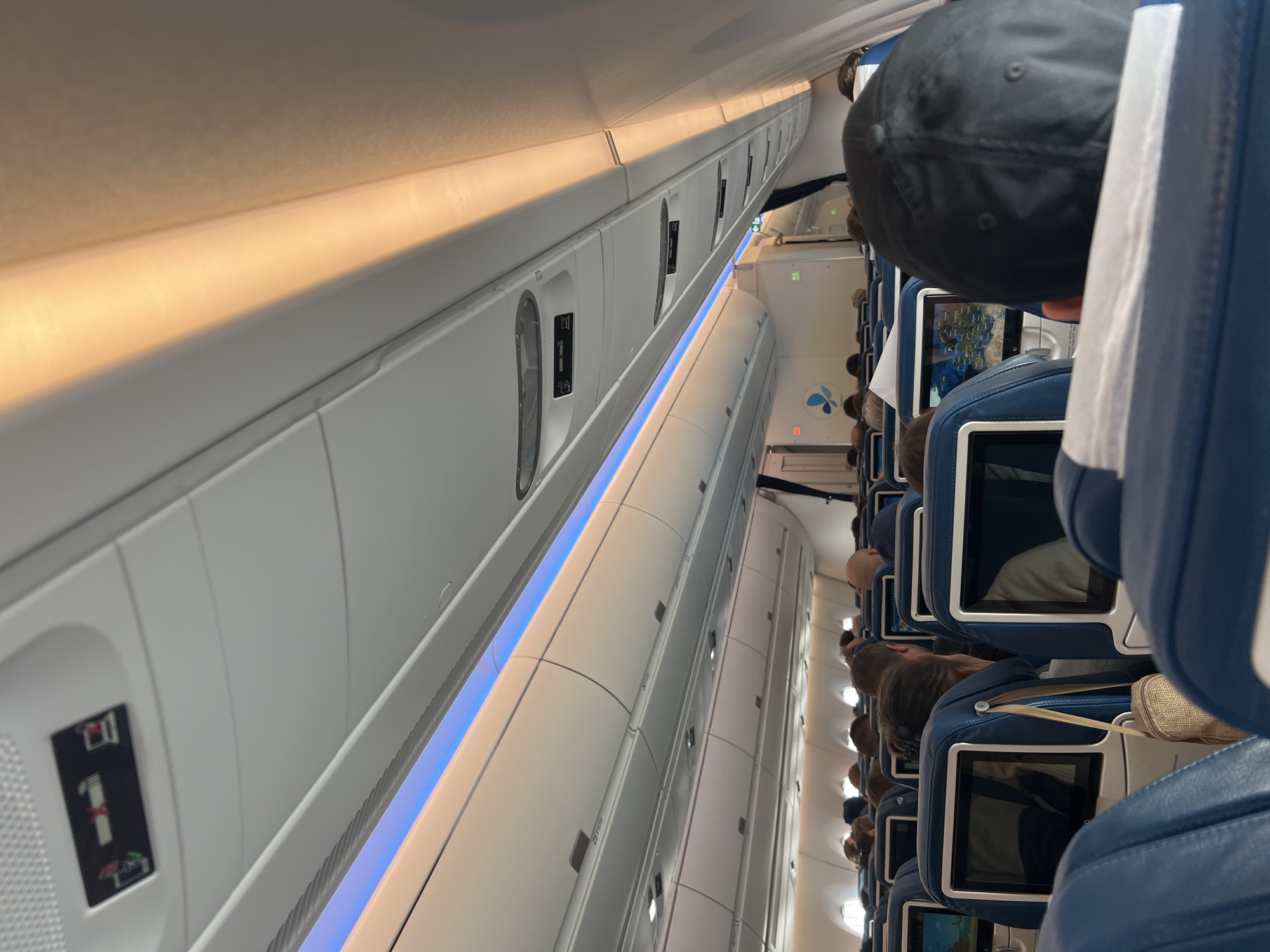 The image shows the interior of an airplane cabin, likely a narrow-body aircraft. Passenger seating is arranged in rows, with the aisle running along the right side of the image. Overhead bins are visible on the left, illuminated by both warm and cool-toned lighting. Passengers are seated, with the back of their heads and the entertainment screens on the seatbacks visible. The angle of the image suggests it was taken from a passenger seat. The airline logo is visible on a bulkhead further down the aisle. There is no information in the picture connecting the scene with the city of Orly, France.