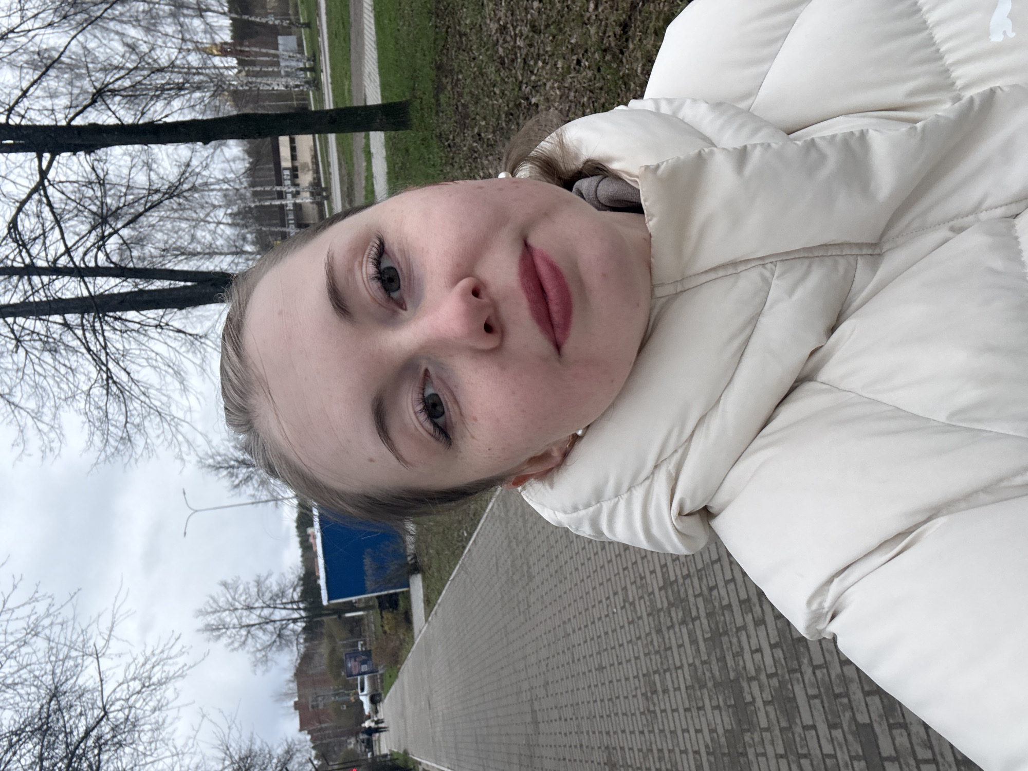 A young woman poses in a selfie. She is wearing a white puffer jacket. The background suggests an urban environment; a brick walkway is visible, along with bare trees, buildings, street signs, and a road with vehicles. The city and precise location are not evident from the photograph.