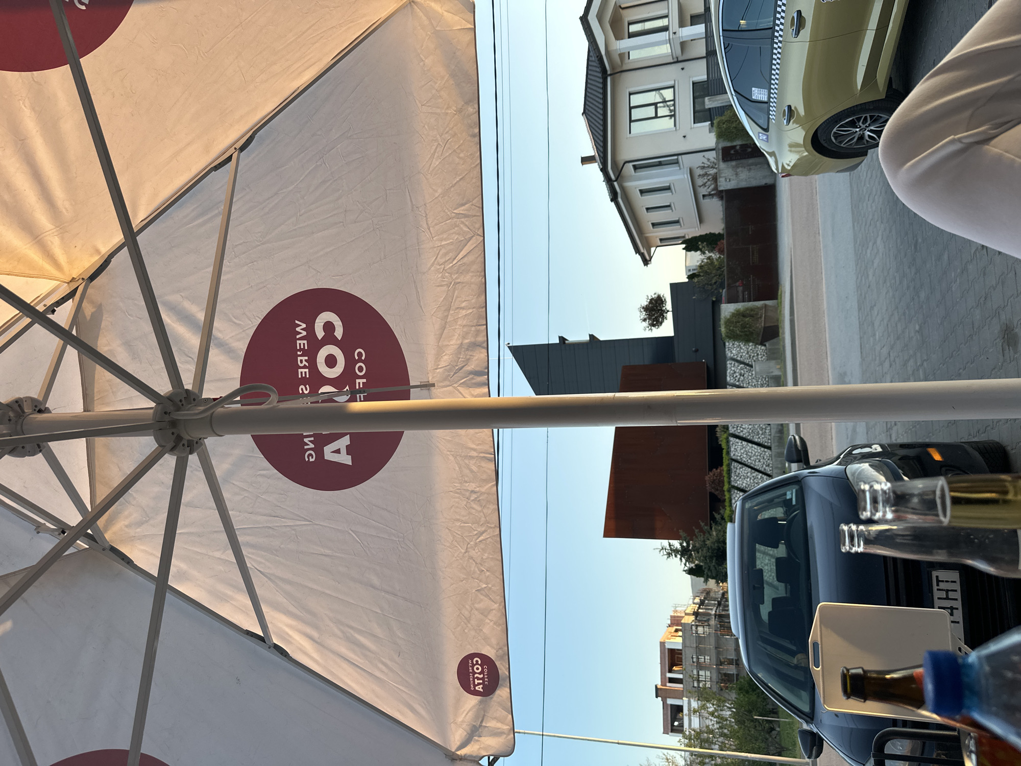 The image shows an outdoor cafe scene in Plovdiv, Bulgaria. A Costa Coffee umbrella dominates the foreground, with the cafe's branding visible. In the background, buildings are visible, including a modern house with a black roof and a traditional-style house with multiple windows. A light-colored taxi is parked in front of the traditional house. A dark-colored car is parked closer to the cafe seating. Bottles and a white serving tray are visible on a table. A person's arm and shoulder are visible on the right edge of the frame. Power lines run across the top of the frame. The sky is clear and blue.