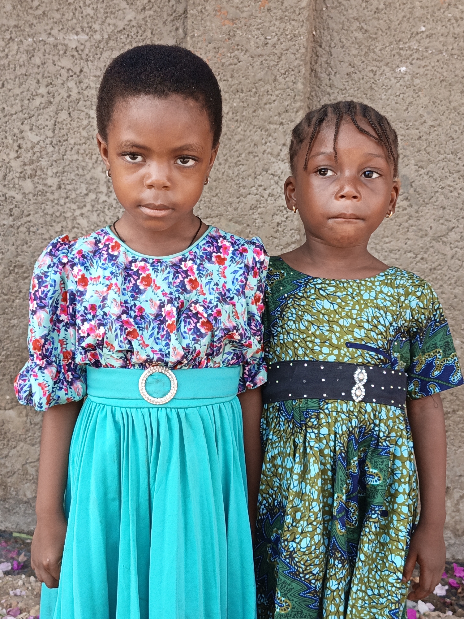 Two young girls stand close together in front of a textured, neutral-toned wall in Gawuna, Nigeria. The girl on the left wears a floral patterned top with puffy sleeves, a solid turquoise belt with a decorative buckle, and a turquoise skirt. The girl on the right wears a patterned dress in shades of green and blue, with a black belt decorated with white dots and a decorative buckle. Both girls have earrings. The setting appears to be outdoors, with some scattered debris or natural material visible near the ground.