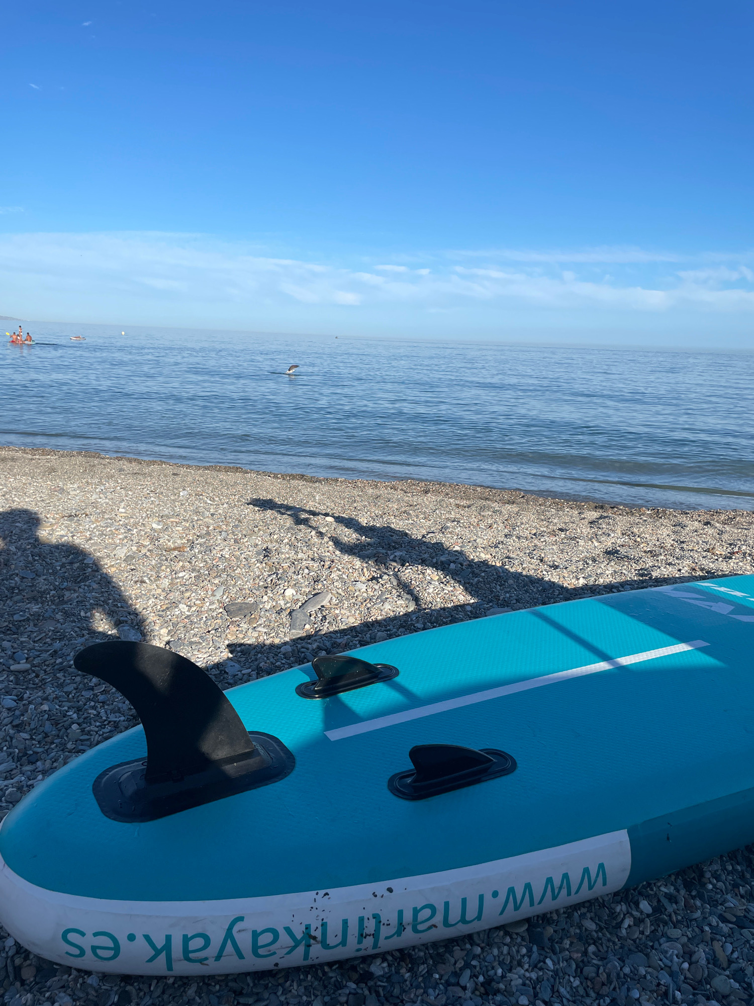 The image shows a blue paddleboard on a pebble beach next to the sea. The paddleboard has two fins and the website "www.martinkayak.es" printed on its side. In the background, the sea is calm, and there are several people on kayaks or paddleboards in the water. A bird is in flight over the water. The sky is clear and blue, with a few wispy clouds. The location is the beach of Lújar, Spain.