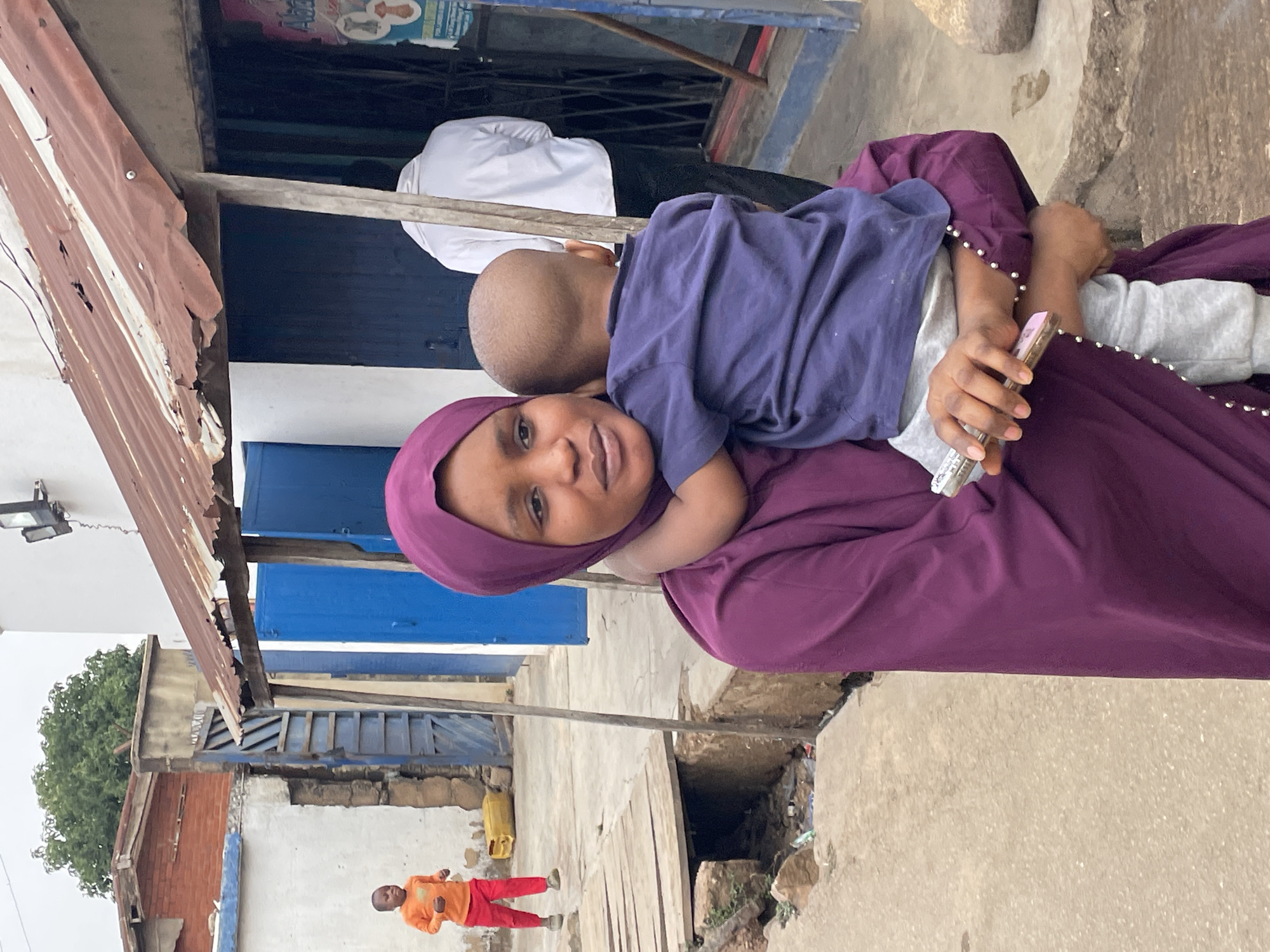 In Jos, Nigeria, a woman wearing a purple hijab and dress is holding a young child. The child wears a blue shirt and grey pants. The woman is holding a cell phone. In the background is a street with buildings of various colors. Another child is wearing an orange shirt and red pants and appears to be walking. The environment suggests a lower-income area.