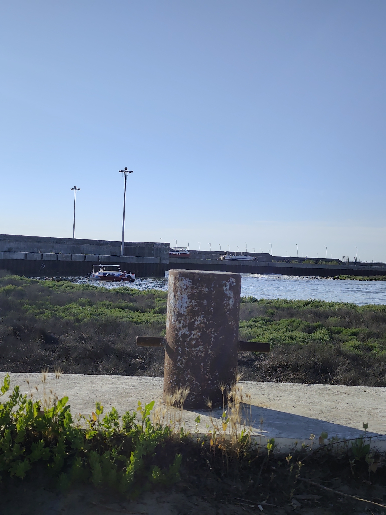 The image shows a concrete embankment with a rusty bollard in the foreground. Beyond the bollard, there is a body of water, likely the Caspian Sea, with a concrete seawall running along its edge. Two boats are visible on the water. The sky is clear and blue. In the distance, there are several tall lampposts. The vegetation consists of low-lying green bushes and some dry grasses. This scene is located on the embankment in the city of Aktau, Kazakhstan, facing the Caspian Sea.