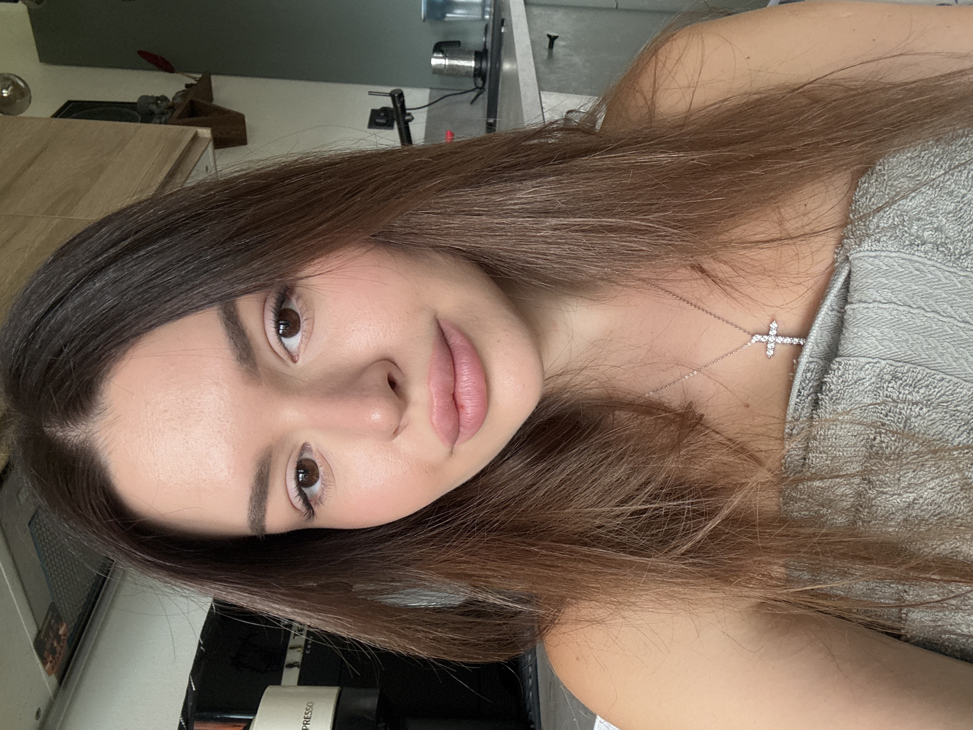 The image is a close-up selfie of a young woman with long, brown hair. She is wearing a silver cross necklace and is wrapped in a grey towel, suggesting she has recently showered. Her makeup is subtle, with emphasis on her eyes and lips. The background appears to be a kitchen, with cabinets, a coffee machine, and a kettle visible. The lighting is natural, coming from a window or overhead source. There are no explicit indicators in the image that place the location in Prague, Czechia.