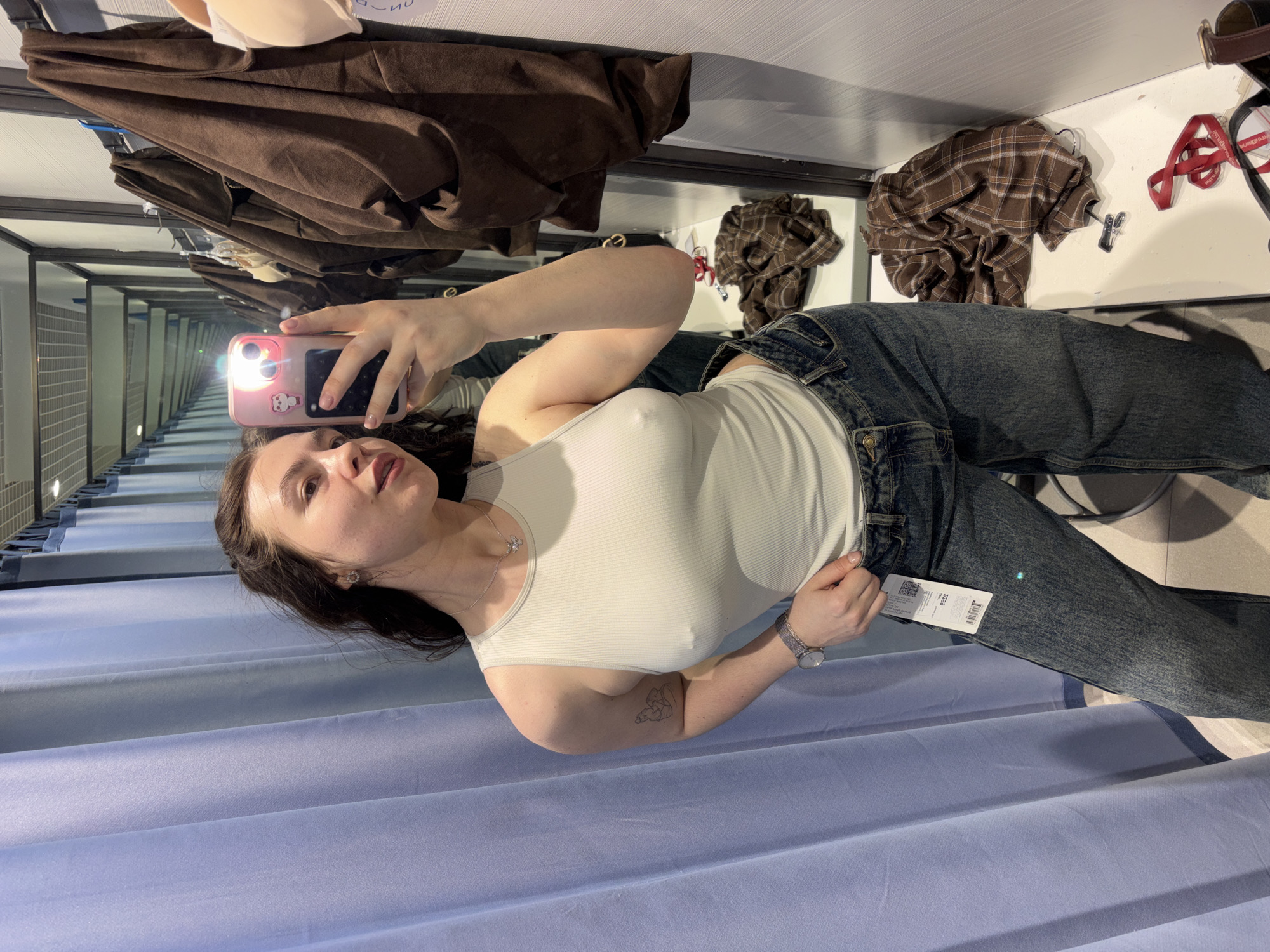 A young woman is captured in a mirror selfie within what appears to be a fitting room, likely in a clothing store in Perm, Russia. She has dark hair and is wearing a ribbed white tank top and blue jeans. Her left hand holds a pink smartphone with a light-colored case, angled to take the picture, while her right hand rests on the waistband of her jeans. A small, outline tattoo is visible on her right forearm, and she wears a silver necklace and a watch on her left wrist.
The setting is an indoor changing room, characterized by blue privacy curtains on one side and a large mirror reflecting the scene. This mirror also reflects the opposite wall, which is lined with more mirrors and changing rooms, creating an optical illusion of an endless corridor. Above the mirror, several items of clothing are visible hanging on a rail, including a brown jacket and a brown plaid shirt. A clothing tag, partially visible on the jeans she is wearing, displays the number '3249' and other small text. The lighting is artificial, typical of a retail environment. The woman's expression is neutral, as she poses for the selfie.