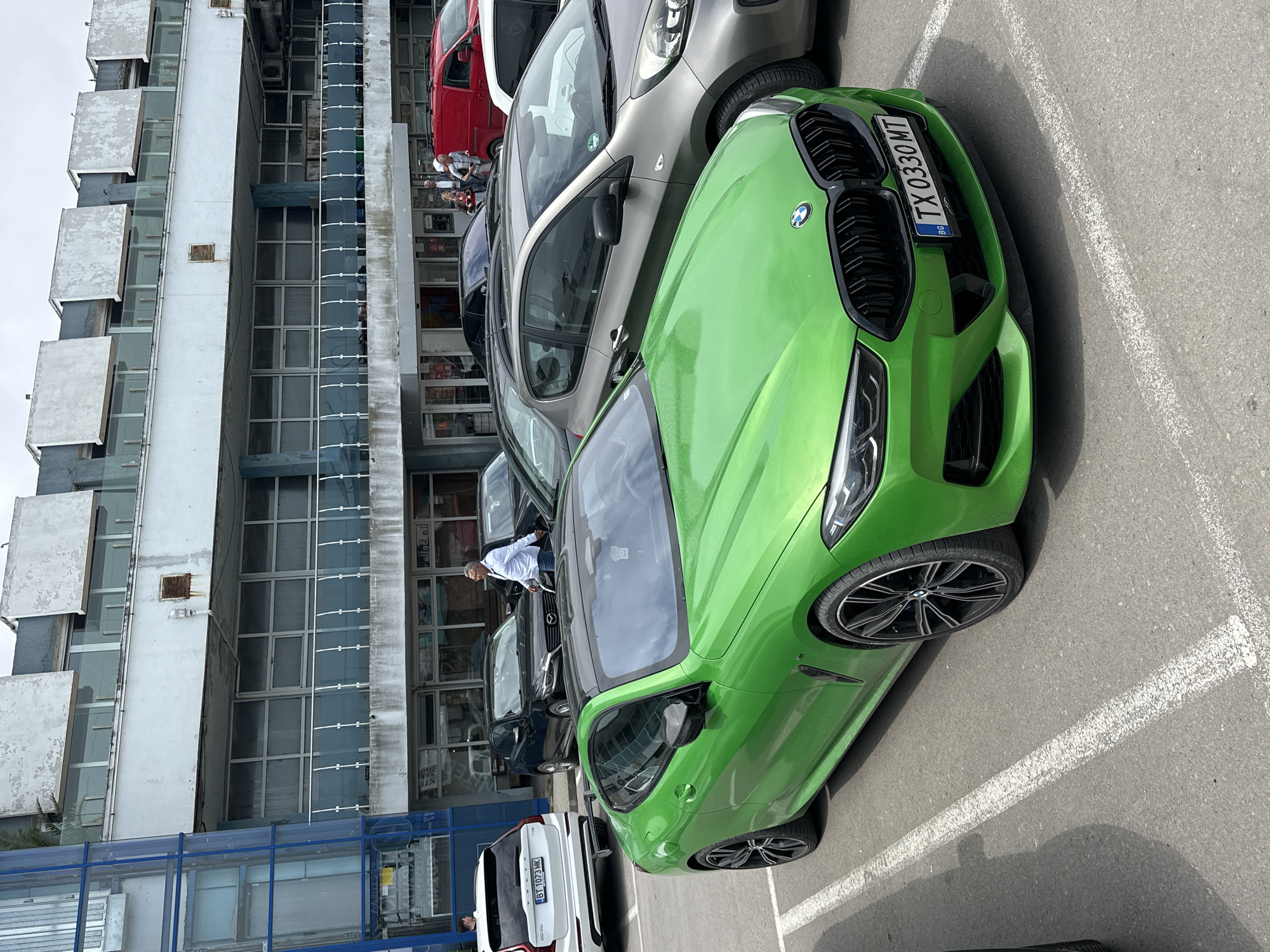 An outdoor scene captures a bustling parking lot adjacent to a multi-story building, likely under a bright, overcast sky during the day. The foreground is dominated by a striking bright green BMW, likely an M-series, with its front facing the viewer. Its large kidney grille and sleek headlights are prominent, along with a visible Bulgarian license plate "TX 0330 MT".
Behind the BMW, a queue of other vehicles is parked, including a gray hatchback (resembling a Kia Picanto), a red van, a dark sedan, and a white Volvo SUV with a "BT 1073 MK" license plate in the distance. Several individuals are present in the parking area; one man in a white shirt is seen leaning into the driver's side of the green BMW, while others are gathered near the red van or are partially visible further back.
The multi-story building itself features numerous windows and concrete balconies, some displaying signs of age and wear. A distinctive blue glass structure, possibly an elevated walkway or architectural detail, runs along a section of the building's facade. This urban setting, combined with the Bulgarian license plates, places the scene in Bulgaria, specifically in the region of Varna as per the provided context. The overall atmosphere suggests a typical daily activity in a commercial or residential urban area.