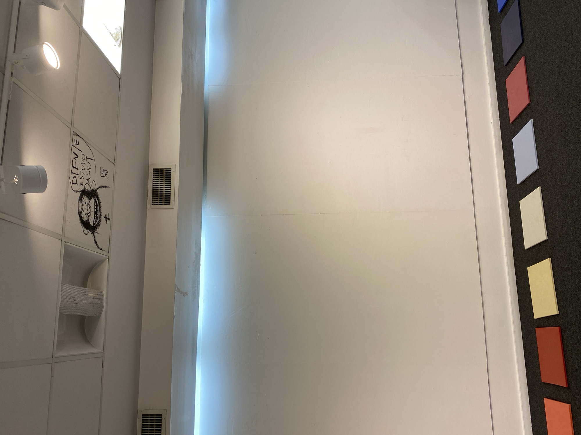 An interior space, likely in Buenos Aires, Argentina, features a white ceiling with various fixtures and a prominent wall on the right. The ceiling is composed of large, rectangular white panels. Integrated into these panels are multiple light sources: at least one bright, circular spotlight and two elongated strip lights that cast a warm glow. Below the spotlight, a modern, white cylindrical light fixture is visible.
Further down the ceiling, a custom-built, white horn-loaded speaker system is recessed into the panels, consisting of two large, flared openings housing a central speaker unit. Next to this, a black graffiti-style drawing is present, depicting a stylized, somewhat grotesque face with large eyes and a wide, grinning mouth. Above this drawing, in black block letters, is the text "DIENTE ESTUVO AQUI," which translates to "DIENTE WAS HERE" (Diente could be a name or "tooth"). A smaller, simpler drawing of a four-legged creature and a hashtag symbol ("#") are also visible near the text.
Adjacent to the ceiling panels, a vertical white wall features a recessed air vent with a gridded cover. Along the intersection of this wall and another architectural element, a narrow, bright blue LED strip light runs vertically, creating a luminous accent. To the far right, a section of a dark gray, possibly acoustic, panel wall is visible, adorned with several small, rectangular, uniformly sized tiles in various colors arranged vertically. From top to bottom, these include blue, dark gray, red, light blue, cream/yellow, orange, and brown.
The scene is static, showing no human activity. The overall impression is one of a modern, perhaps industrial or artistically inclined, indoor environment with a mix of functional elements and personalized touches. The artificial lighting suggests it could be any time of day.